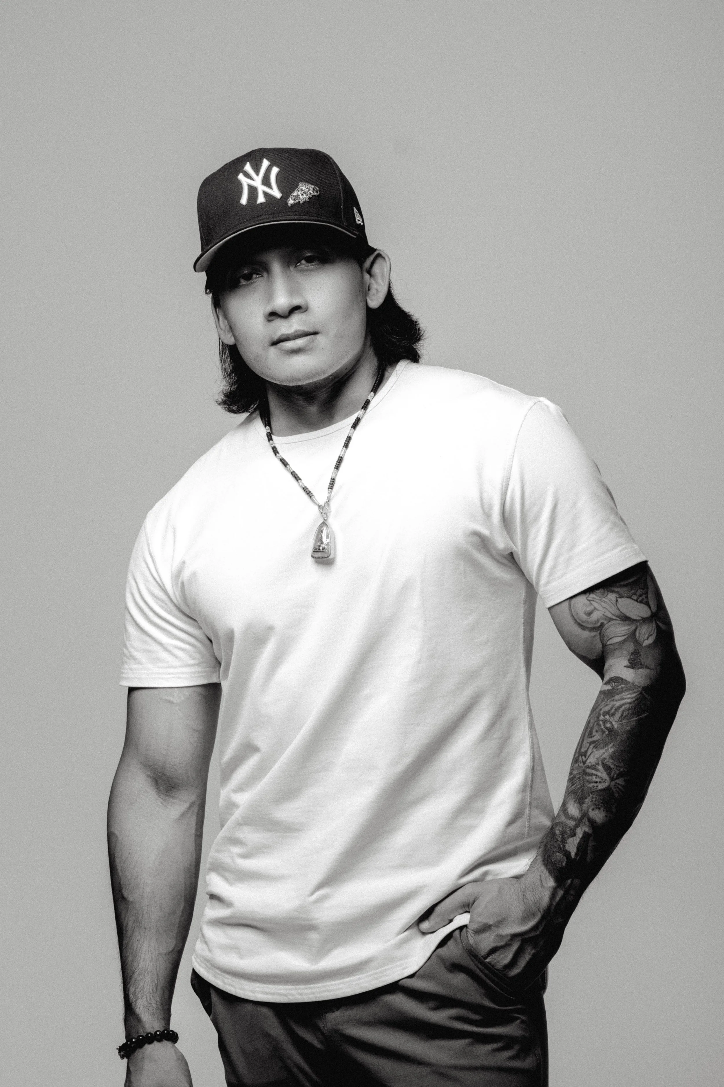 Black and white photo of a young man wearing a baseball cap, t-shirt, necklace, and tattooed arm, standing with one hand in his pocket.