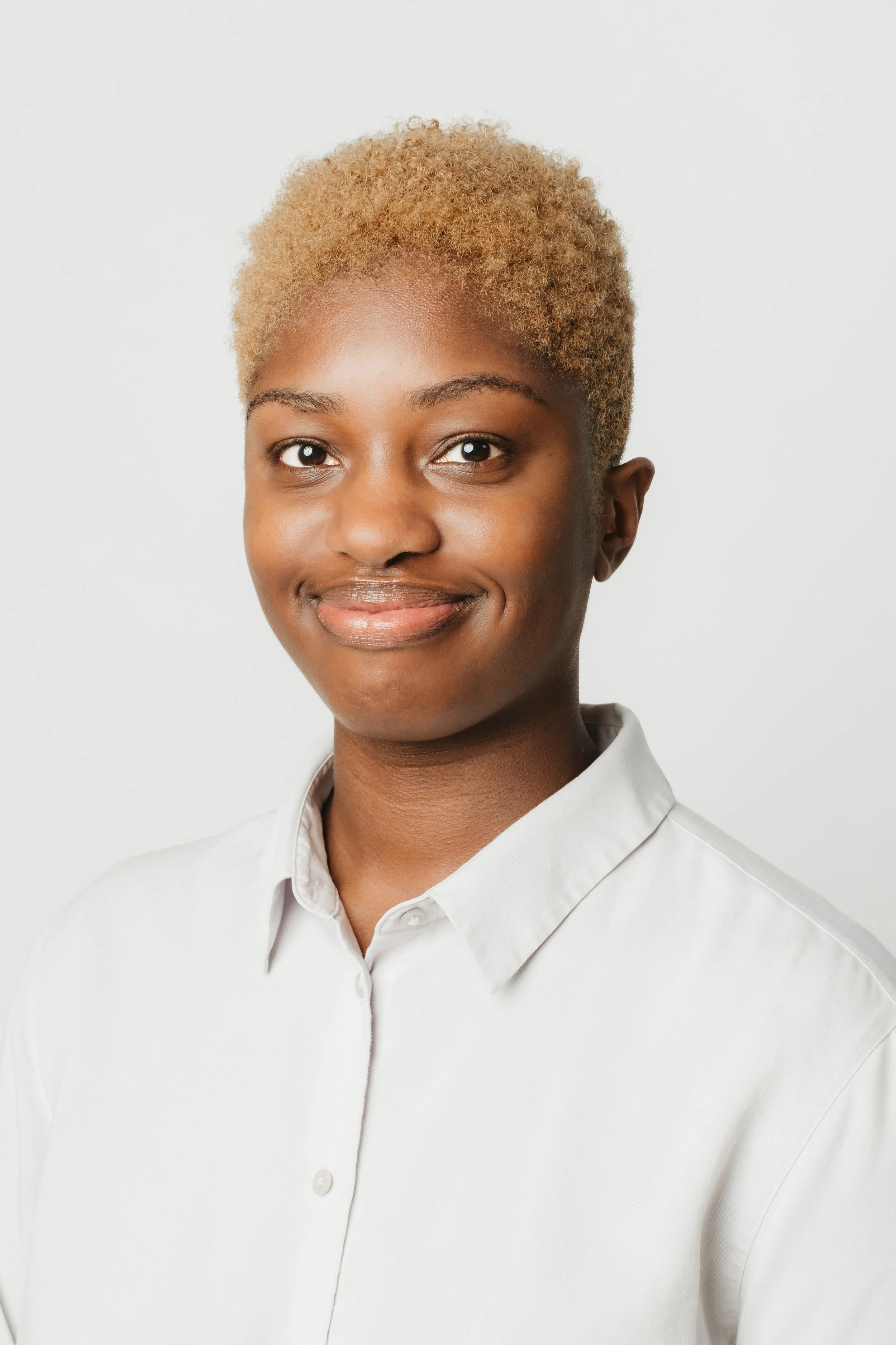 Portrait of a young Black woman with short blond hair wearing a white button-up shirt, smiling softly against a plain white background.