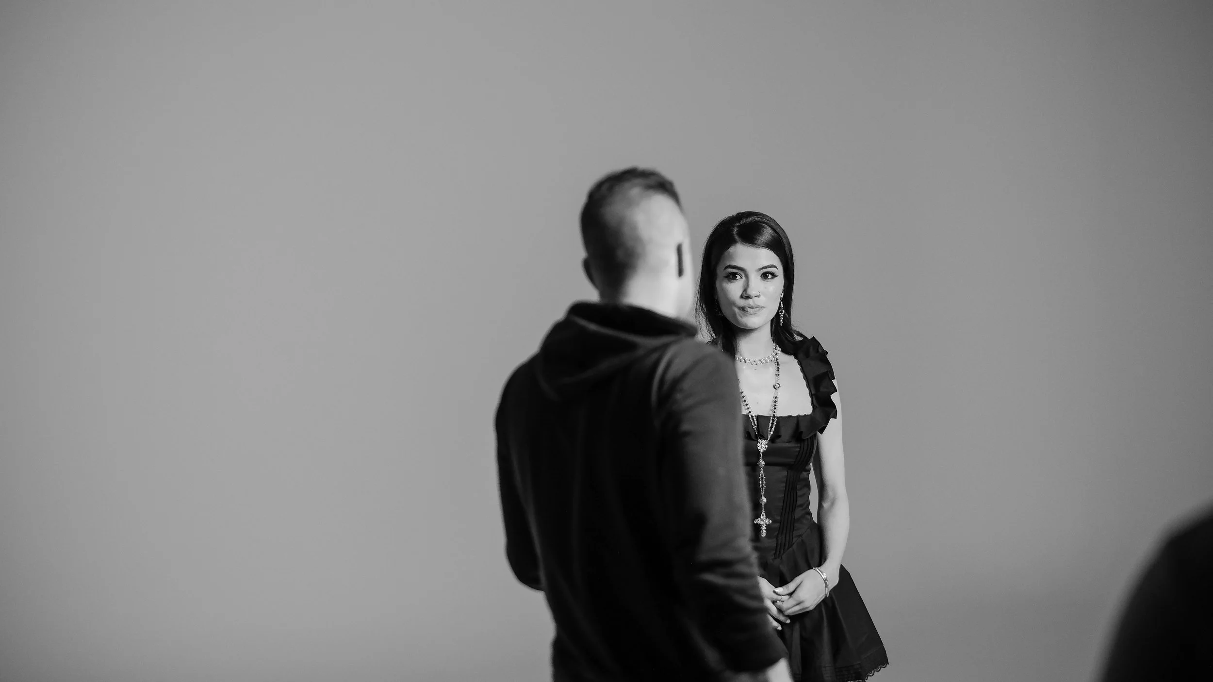 A woman with dark hair, dressed in a black ruffled dress with jewelry, looks at a man with short hair, wearing a dark hoodie, against a plain background.