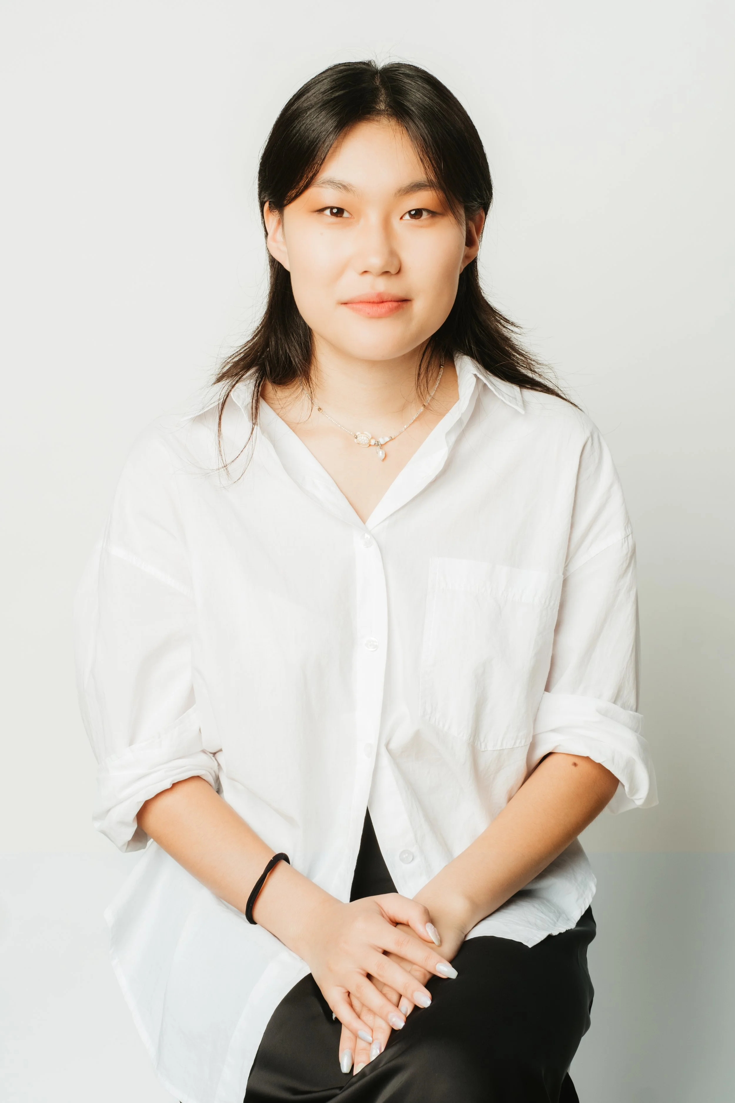 A young woman with black hair wearing a white button-down shirt and black pants, sitting against a plain white background.