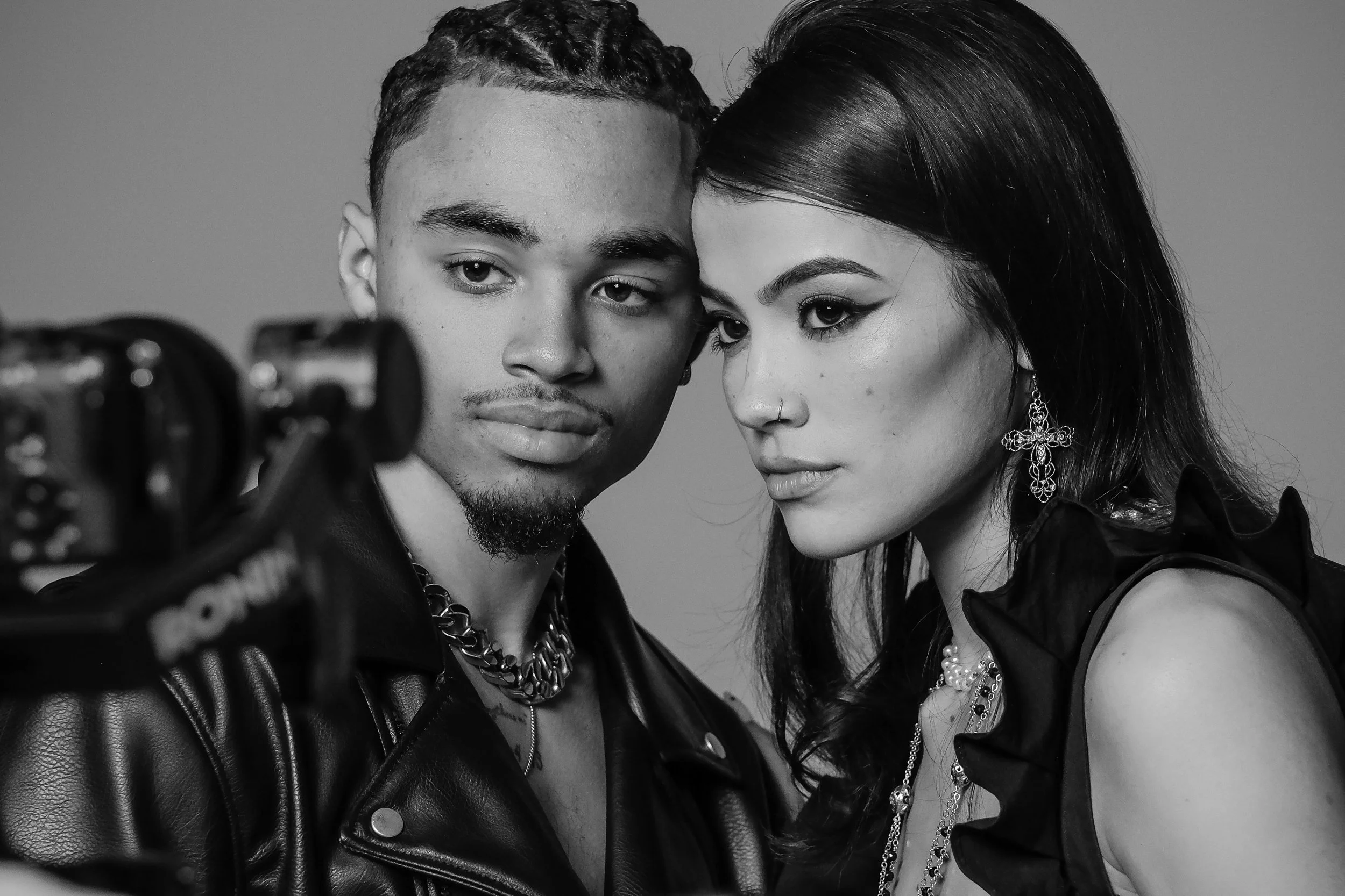 Black and white photo of a man and woman taking a selfie together, both with intense expressions, wearing jewelry and fashionable outfits, with camera visible in the foreground.