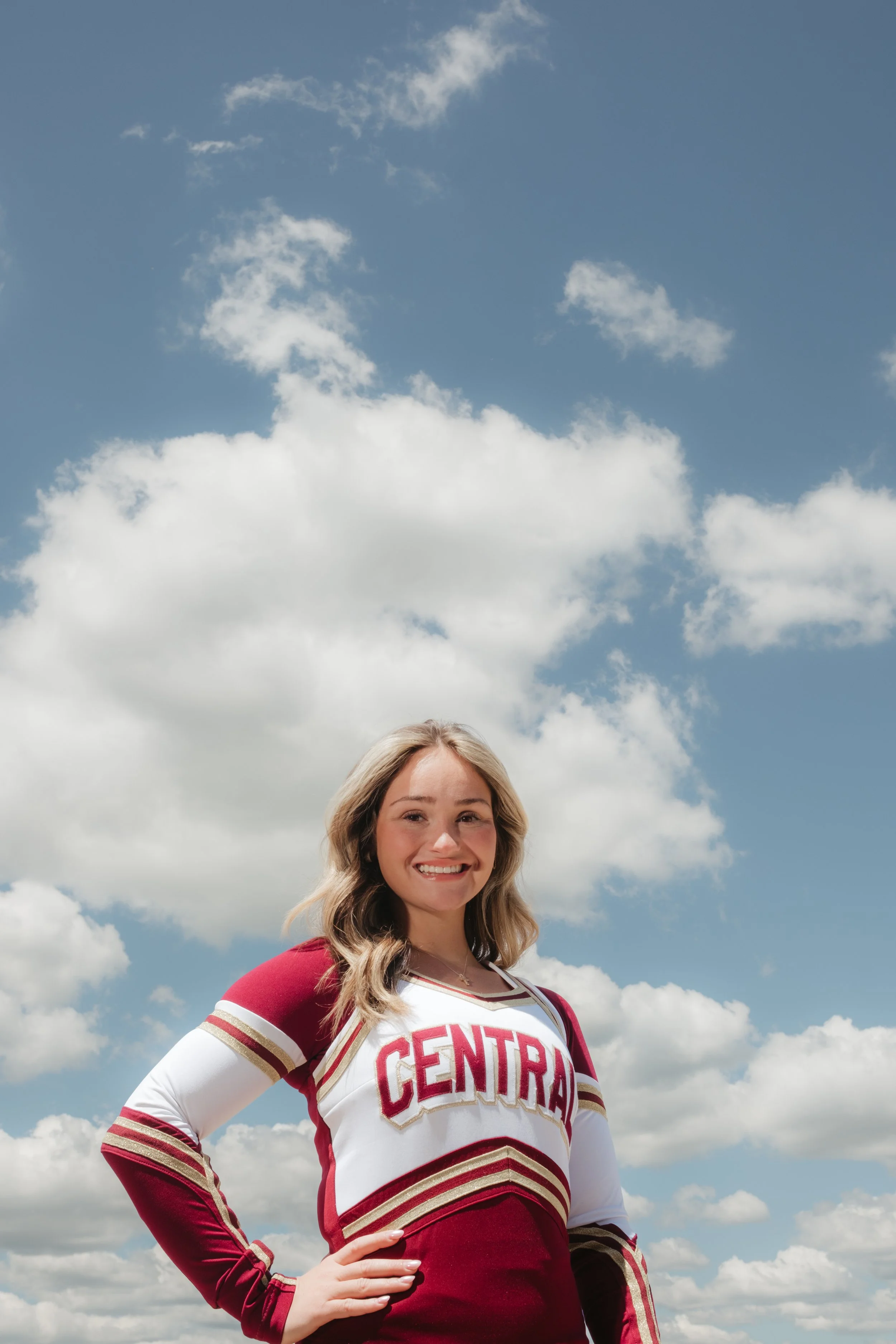 A young woman with blonde hair smiling, standing outdoors against a blue sky with scattered white clouds, wearing a red and white cheerleading uniform with the word 'CENTRAL' on it.