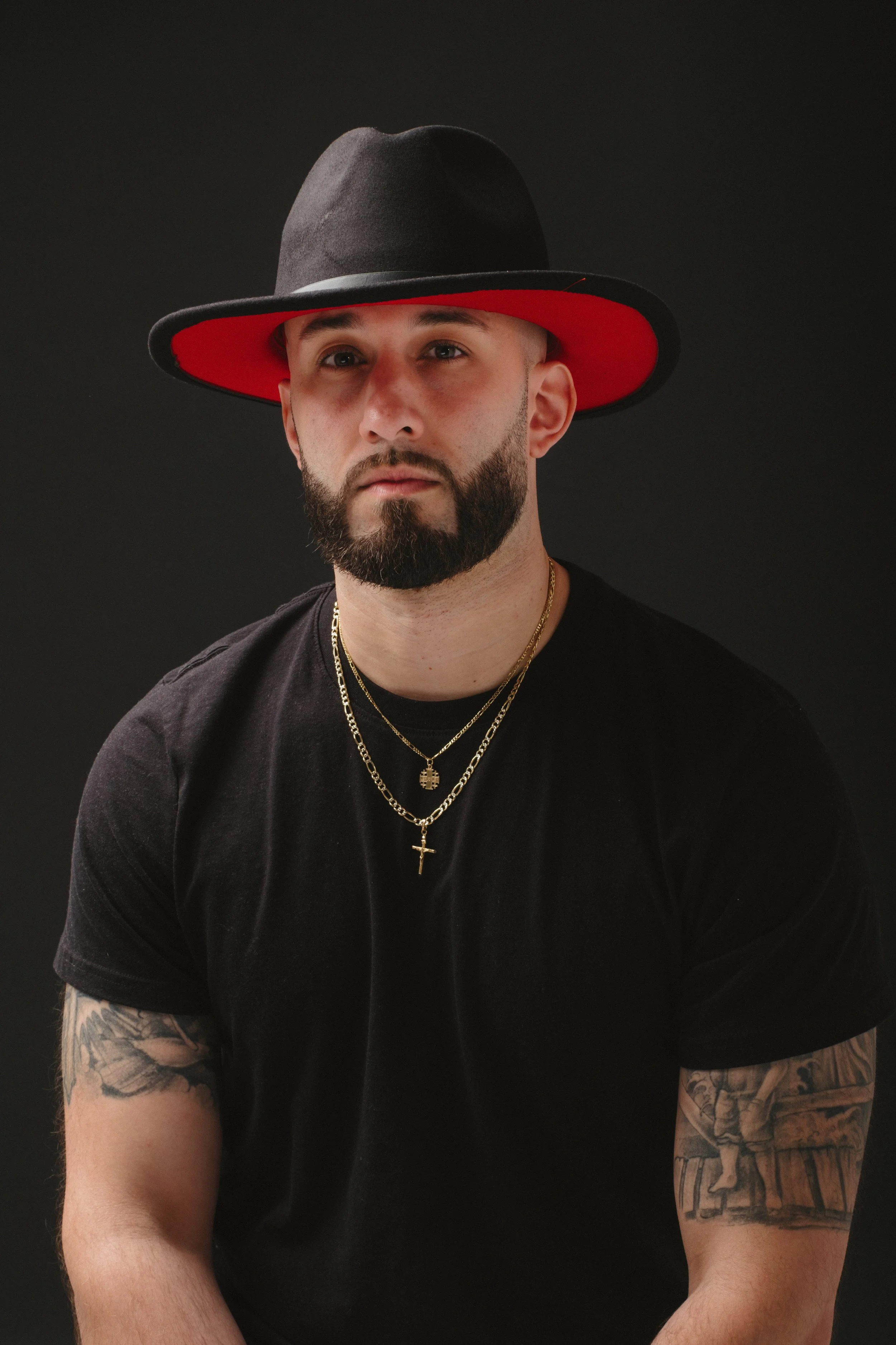 Portrait of a man with a beard wearing a large black hat with a red underside, a black t-shirt, and layered gold necklaces, against a dark background.