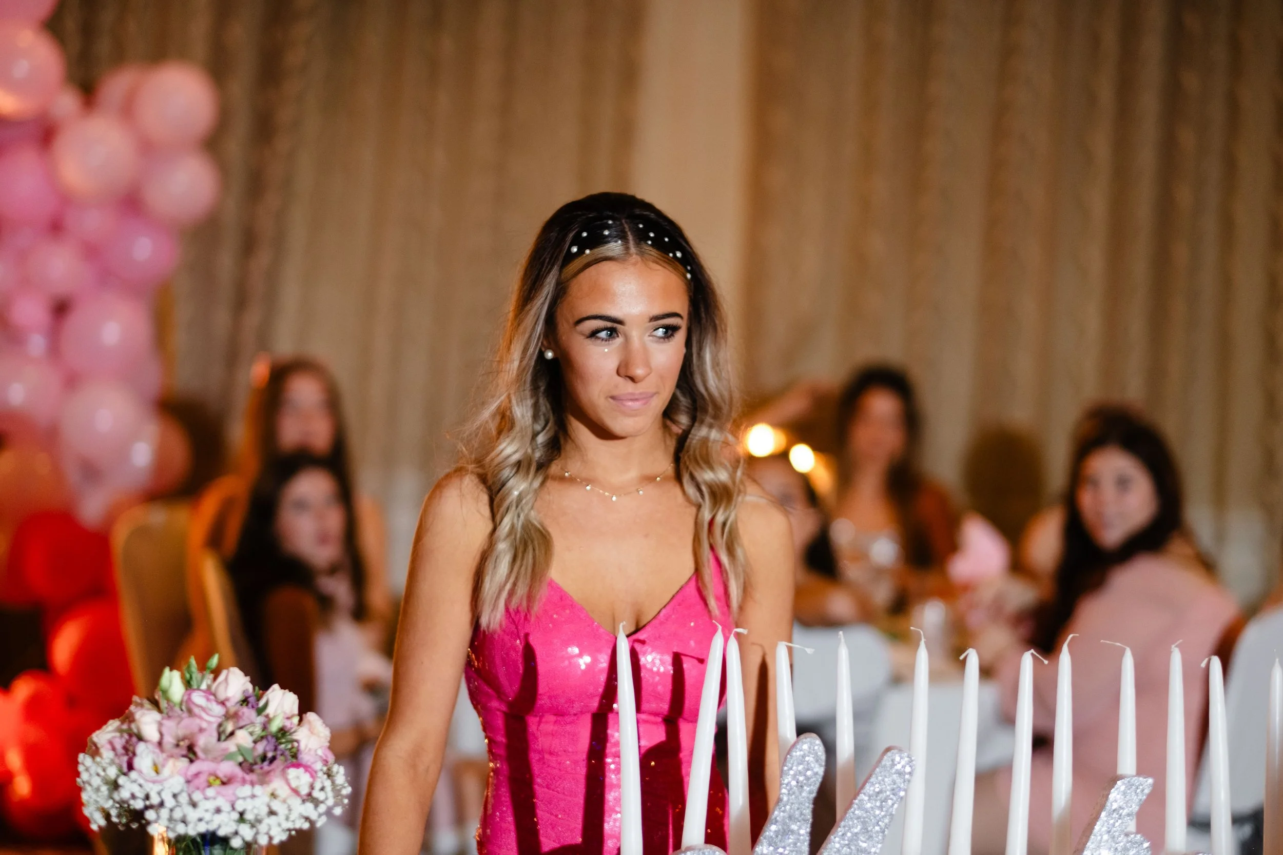 A young woman in a pink dress at a celebration, surrounded by candles and flowers, with other women in the background.
