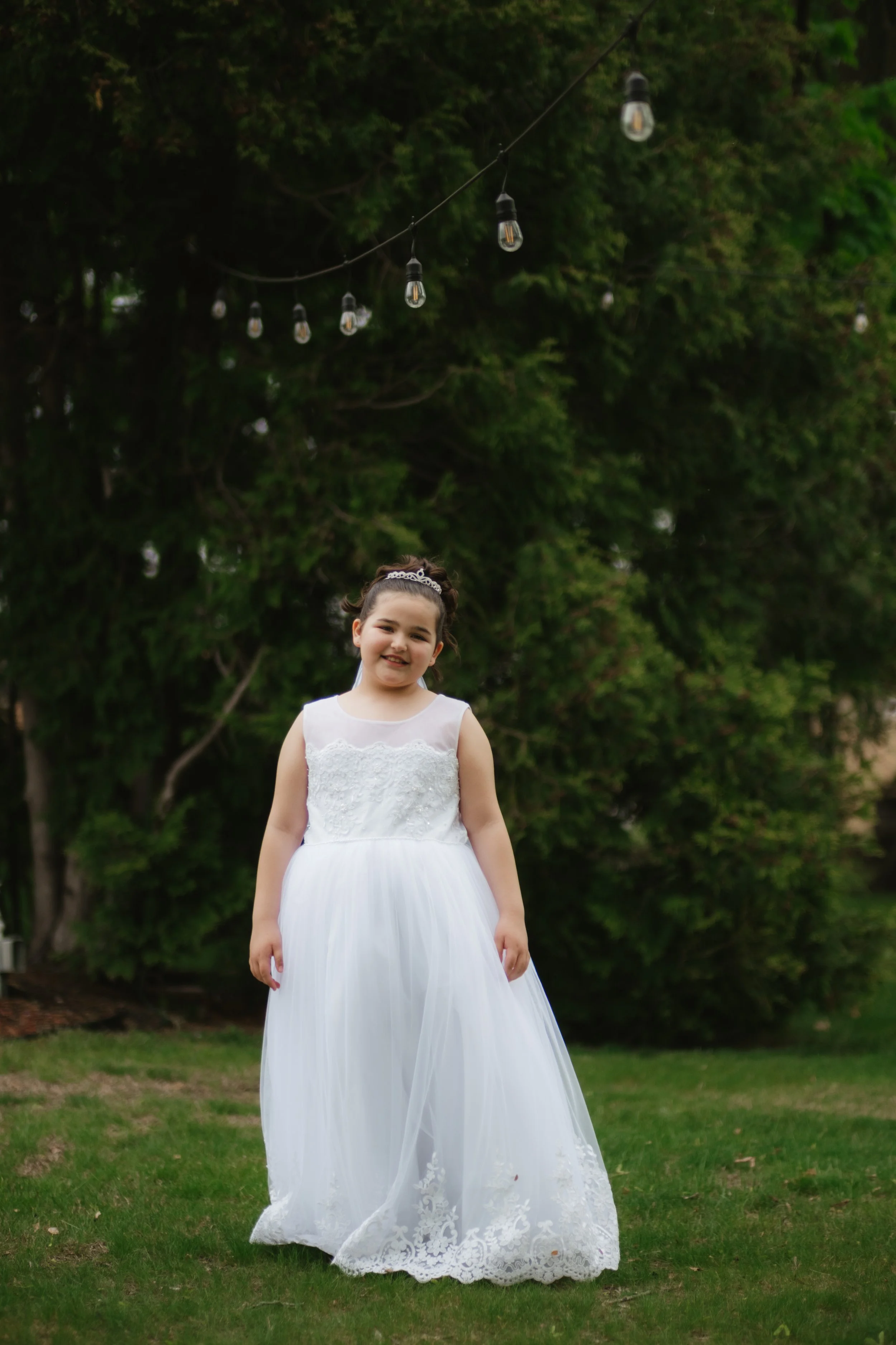 A young girl in a white dress standing on a grassy lawn, with string lights hanging above and green trees in the background.