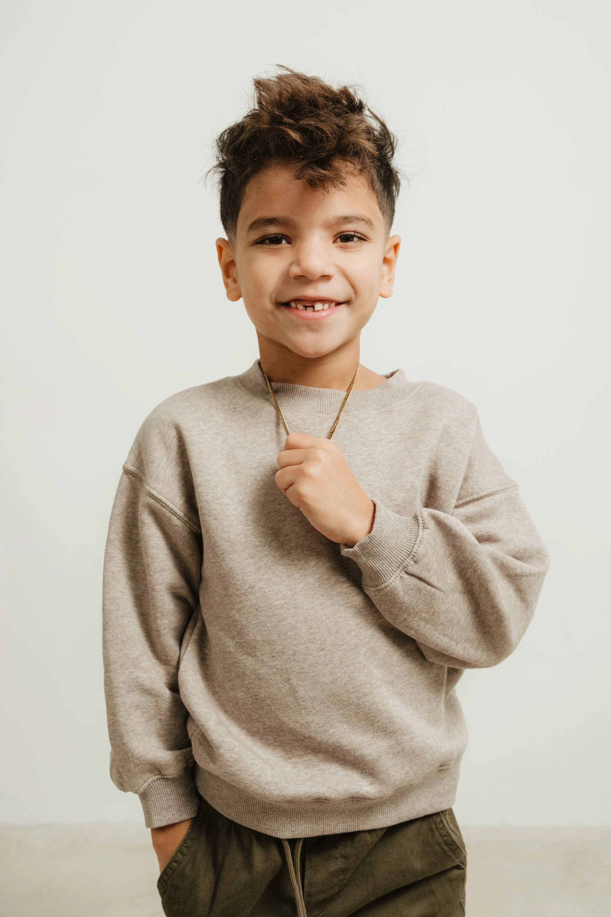 A smiling young boy with curly hair wearing a beige sweatshirt and brown pants, holding a gold chain necklace with his left hand.