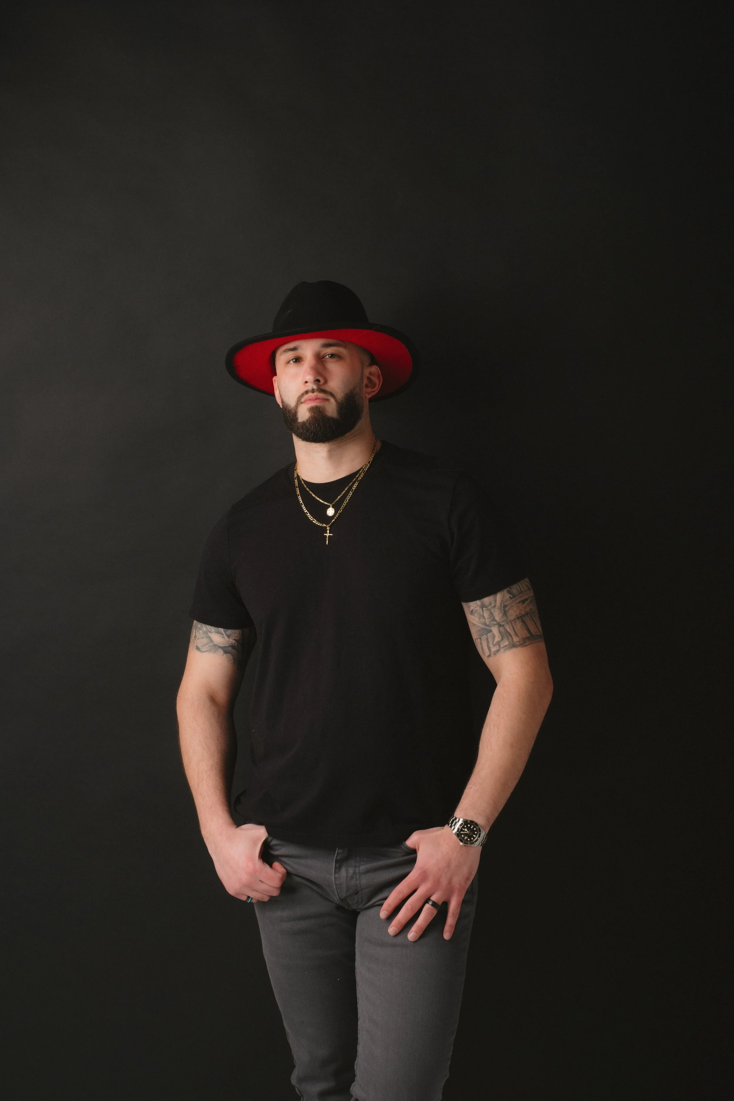 Man wearing a black shirt, gray jeans, gold necklaces, a watch, a ring, and a black hat with a red interior, standing against a black background