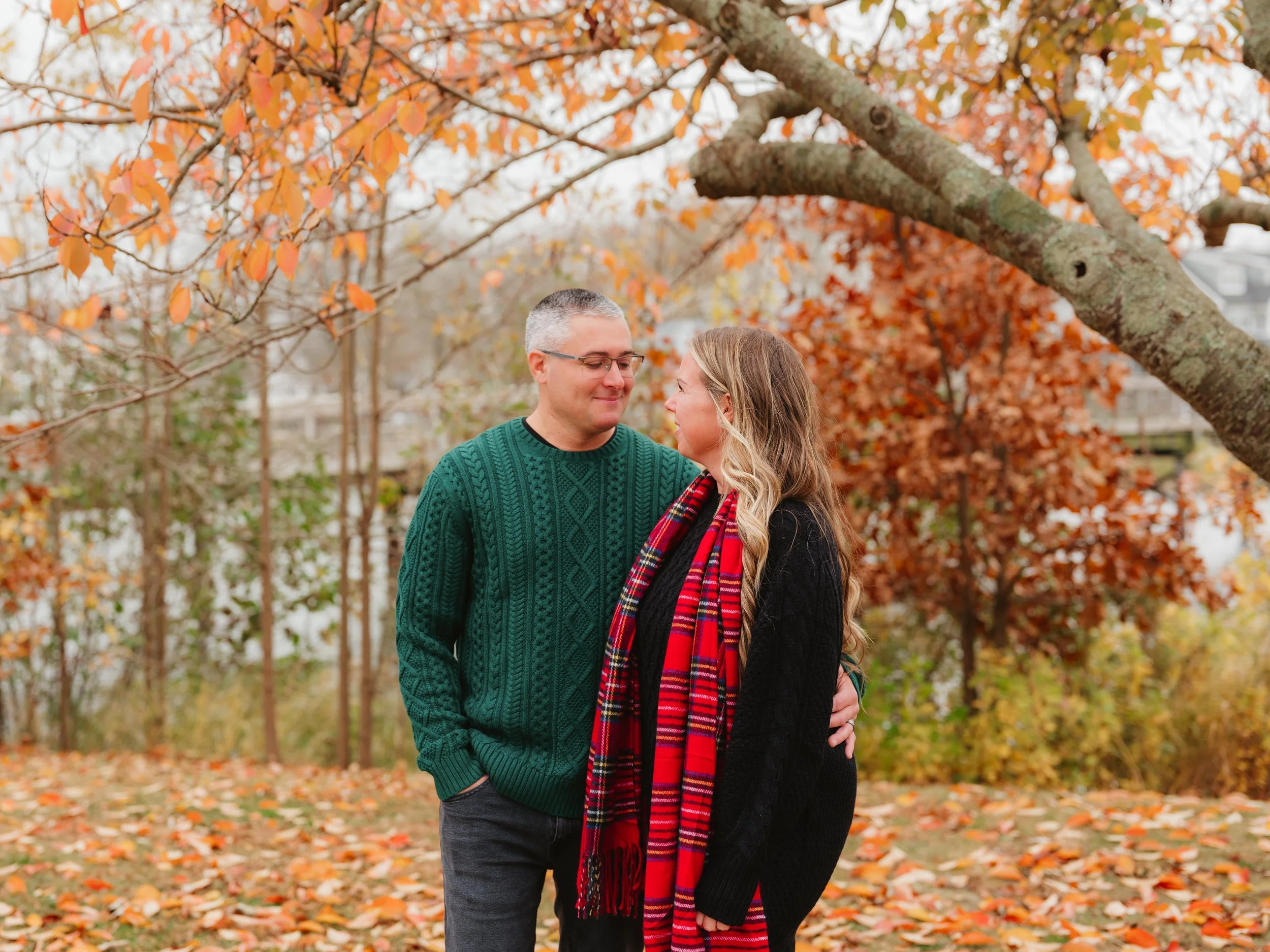 A man and woman standing close together outdoors during fall, under autumn trees with orange leaves, looking at each other affectionately.