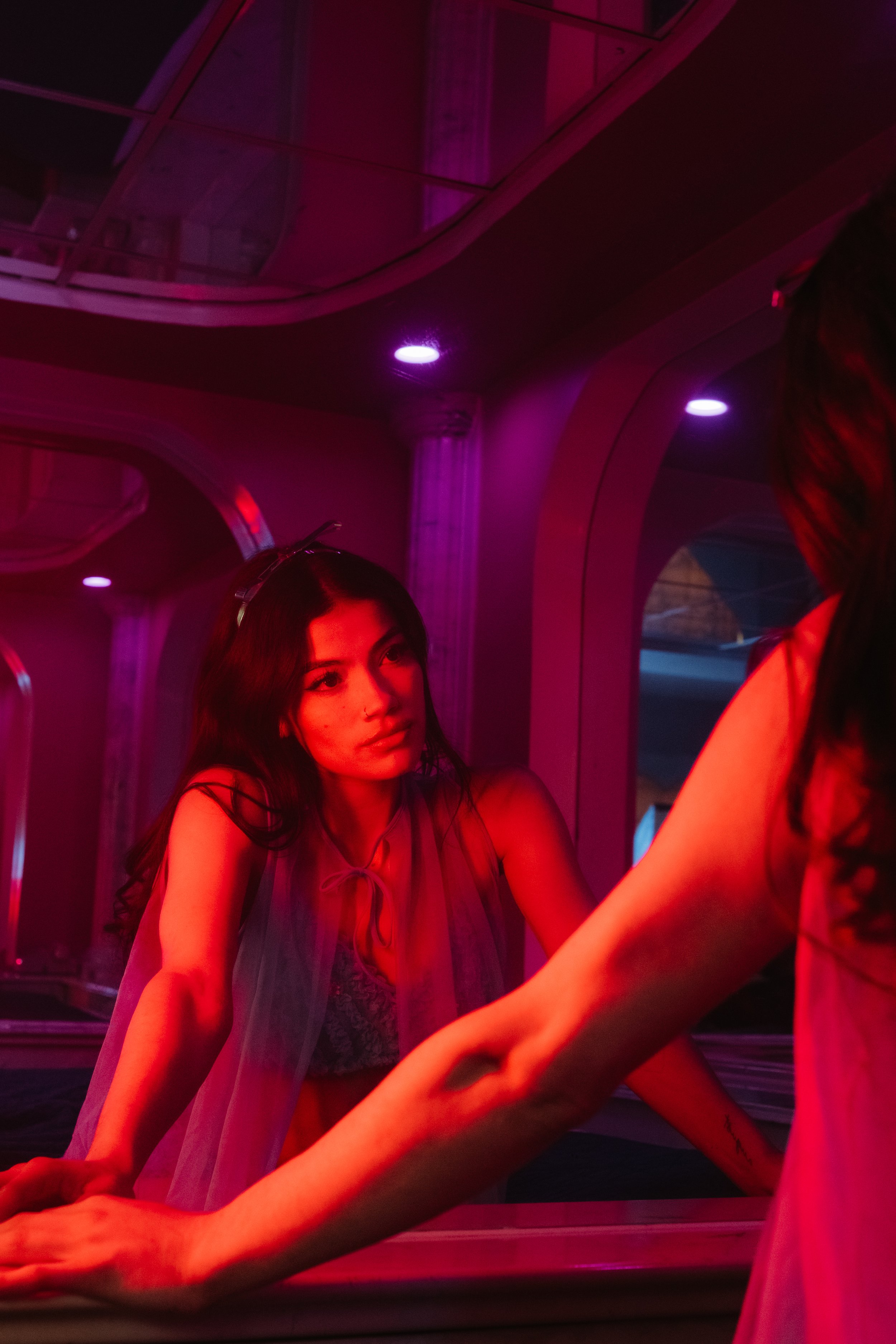 A woman with long dark hair, illuminated by pink and purple neon lights, leaning on a bar counter and looking at another person outside the frame in a dimly lit, colorful club setting.