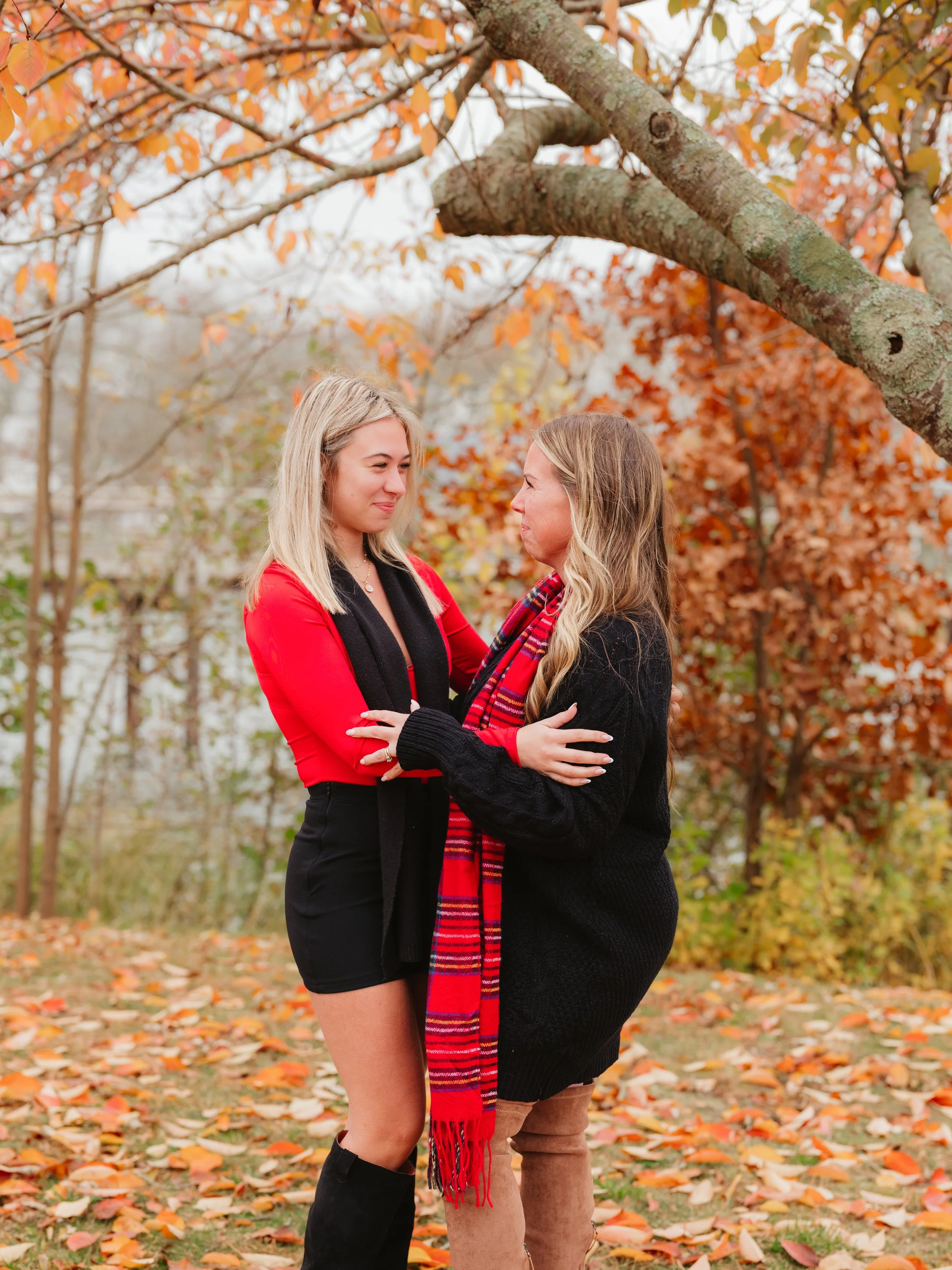 A woman and girl stand facing each other among autumn trees with orange and yellow leaves. They are touching each other's arms and gazing at each other with smiles.
