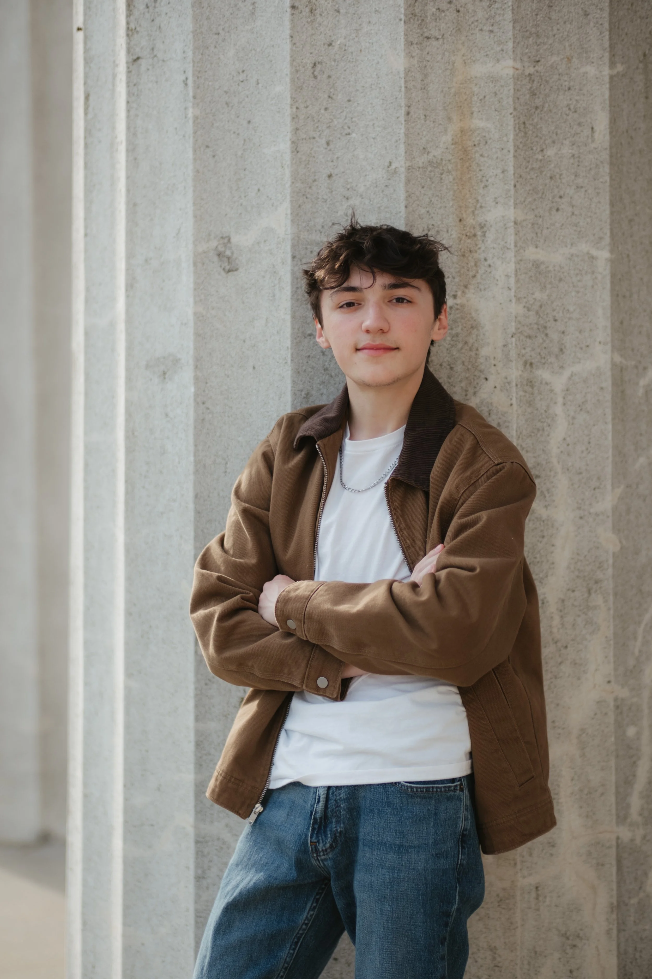 A young man with brown curly hair wearing a brown jacket, white t-shirt, blue jeans, and a silver chain necklace standing with folded arms against a stone wall.