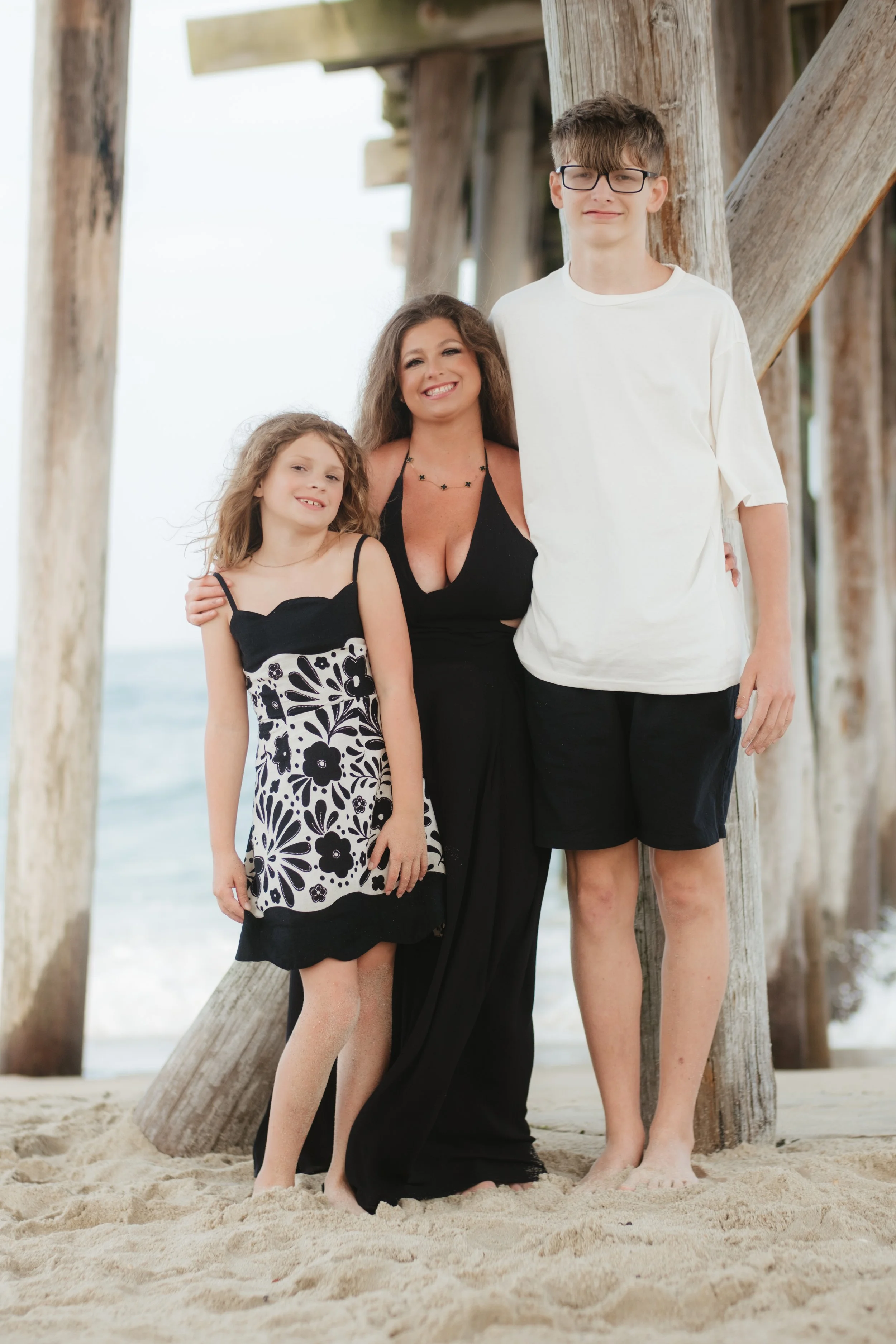 A woman with two children standing under a pier on the beach with ocean in the background.
