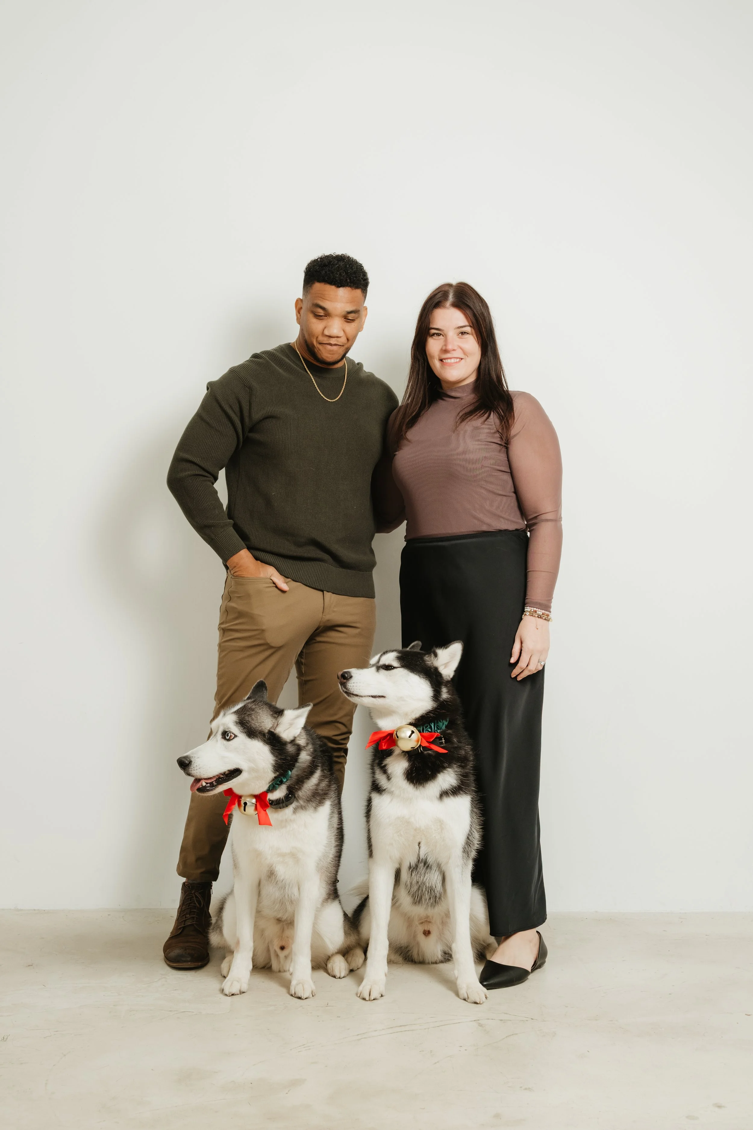 A man and woman stand together with two Siberian Huskies decorated with holiday collars and bells, against a plain white wall.