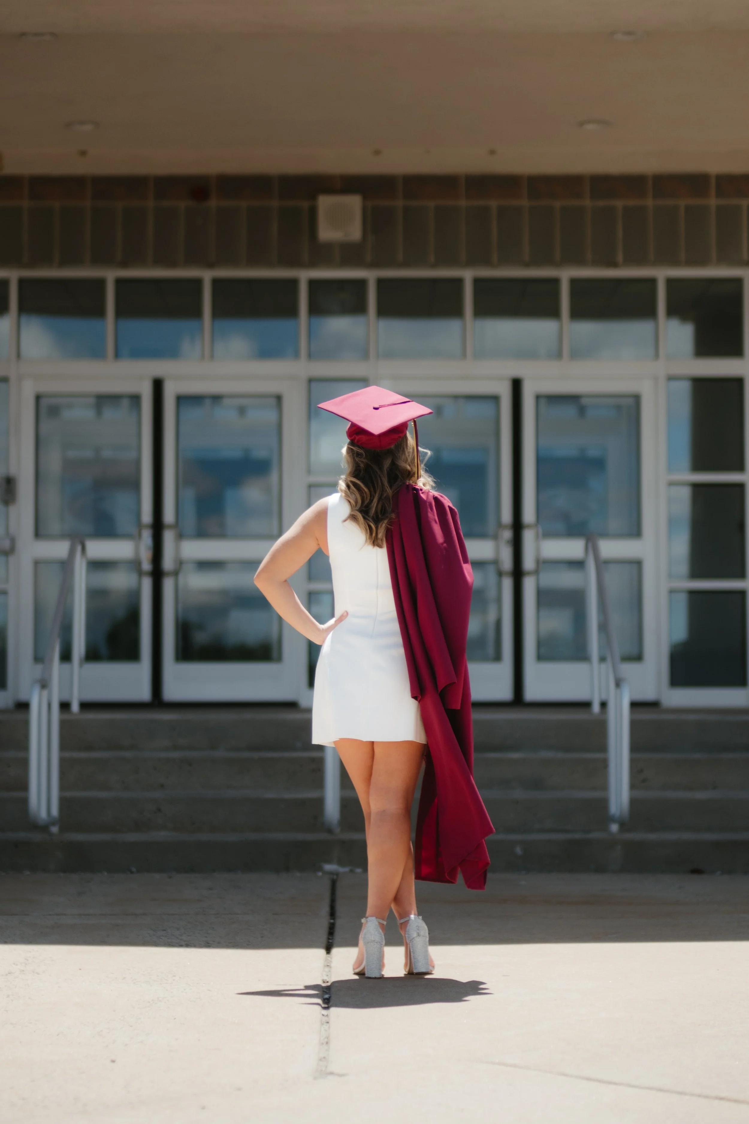 A woman in a white dress, wearing a pink graduation cap and gown, stands outdoors in front of a building with glass doors, with her back to the camera.