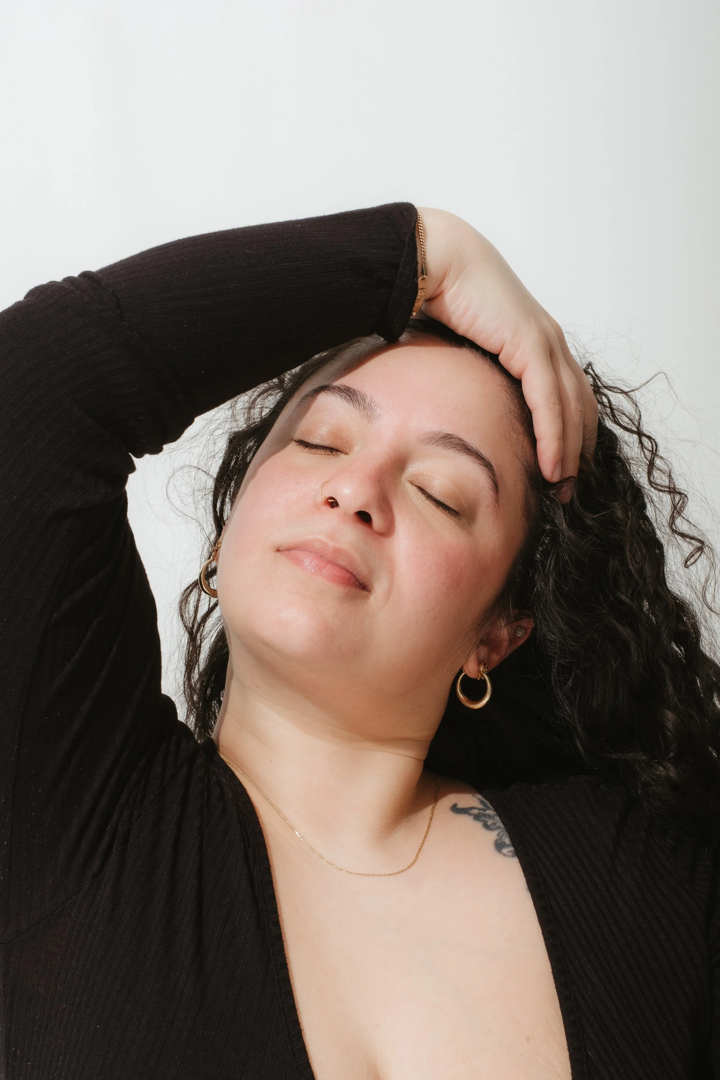 A woman with curly dark hair and light skin, eyes closed, smiling gently, with her hand resting on her head, wearing gold hoop earrings and a black top, against a plain white background.