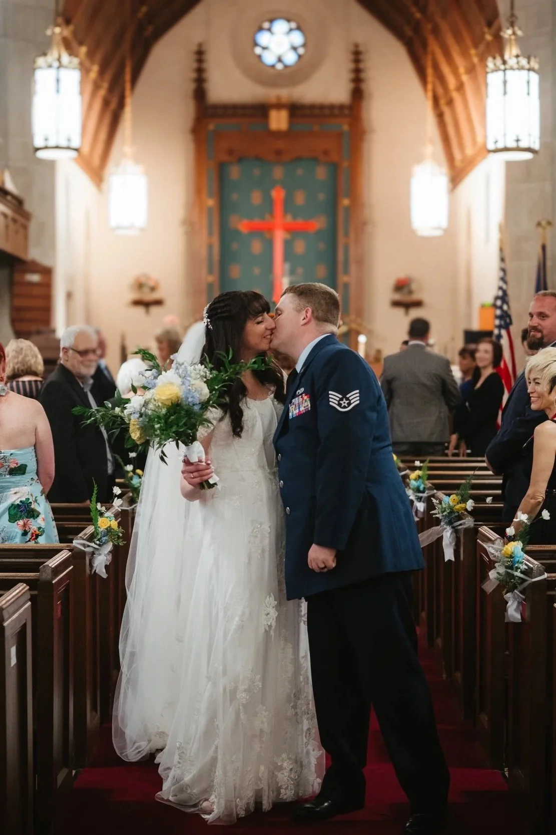 A bride and groom kiss during their wedding ceremony inside a church, with guests seated in pews decorated with flowers and ribbons, and a large red cross visible on the altar background.