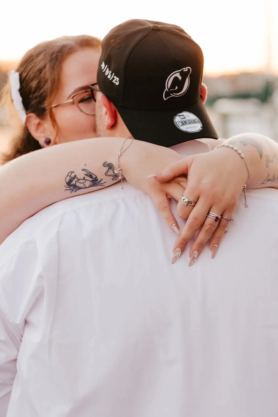 A couple is hugging closely, with one person's arms wrapped around the other's neck, kissing. The woman has tattoos on her arm and fingers, and is wearing rings and bracelets. The man is wearing a black cap and a white shirt.