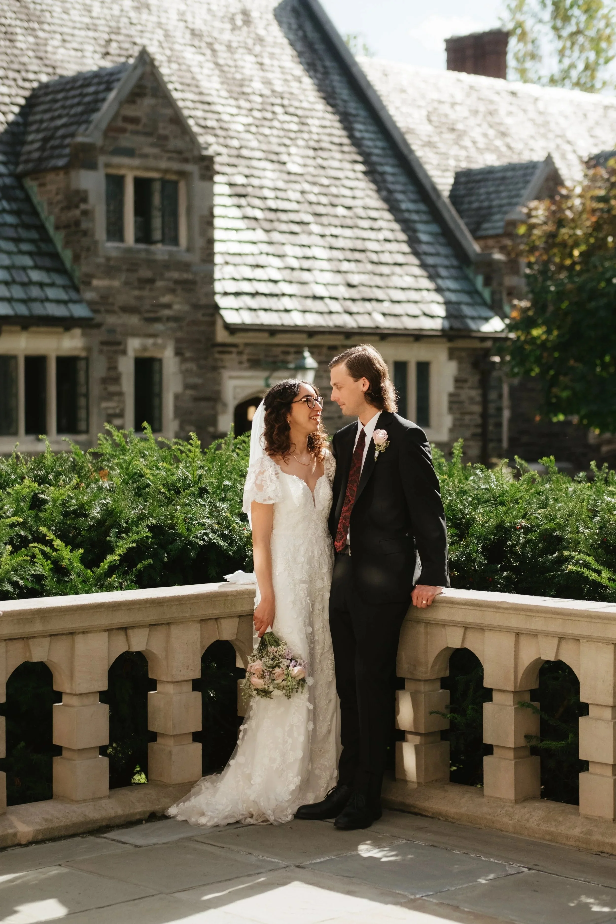 A bride and groom in wedding attire standing close on a stone balcony, smiling at each other with a historic stone house in the background.