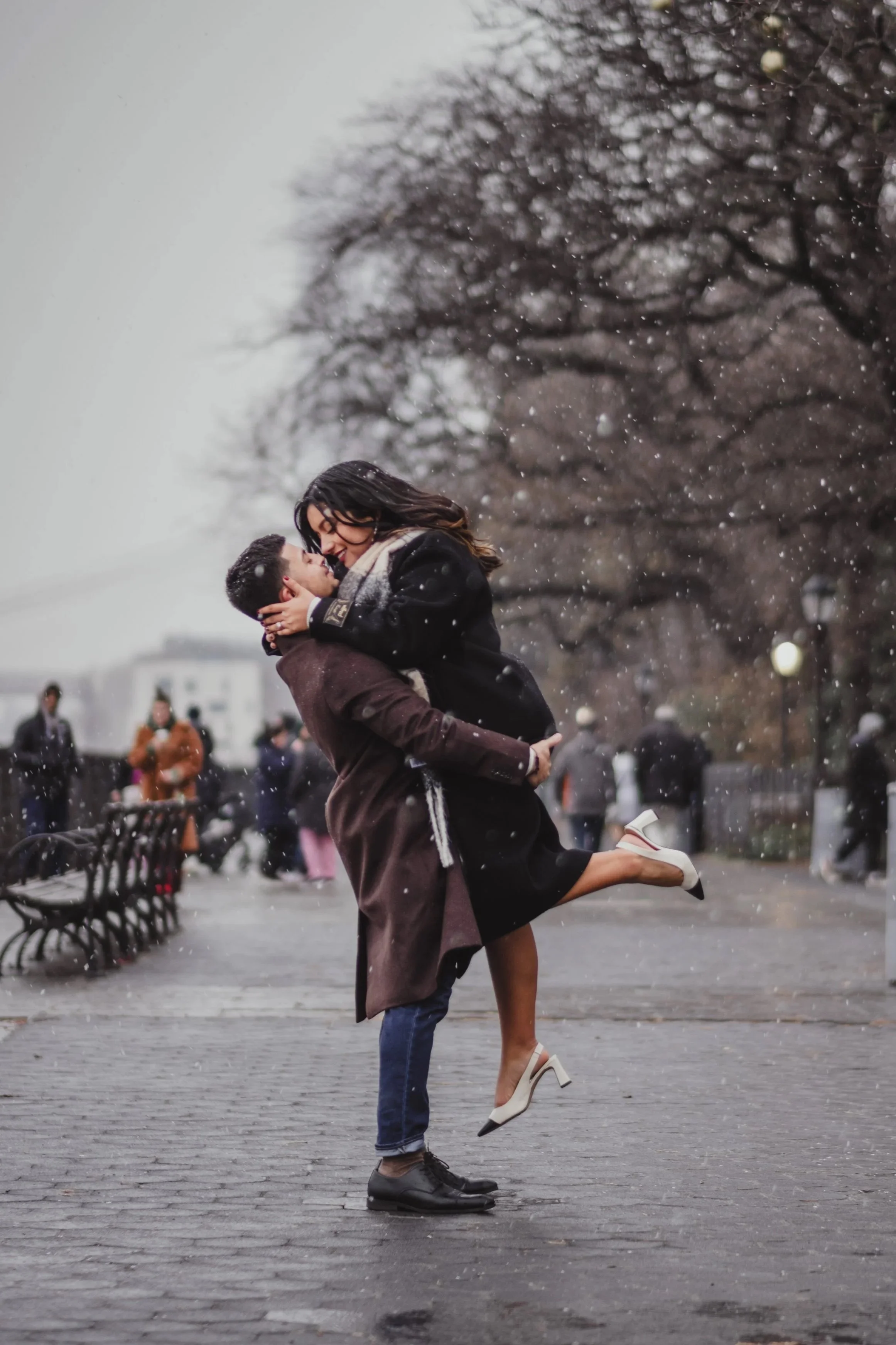 A couple is embracing and lifting each other in a joyful moment outdoors on a rainy day, with other people walking in the background and leafless trees.