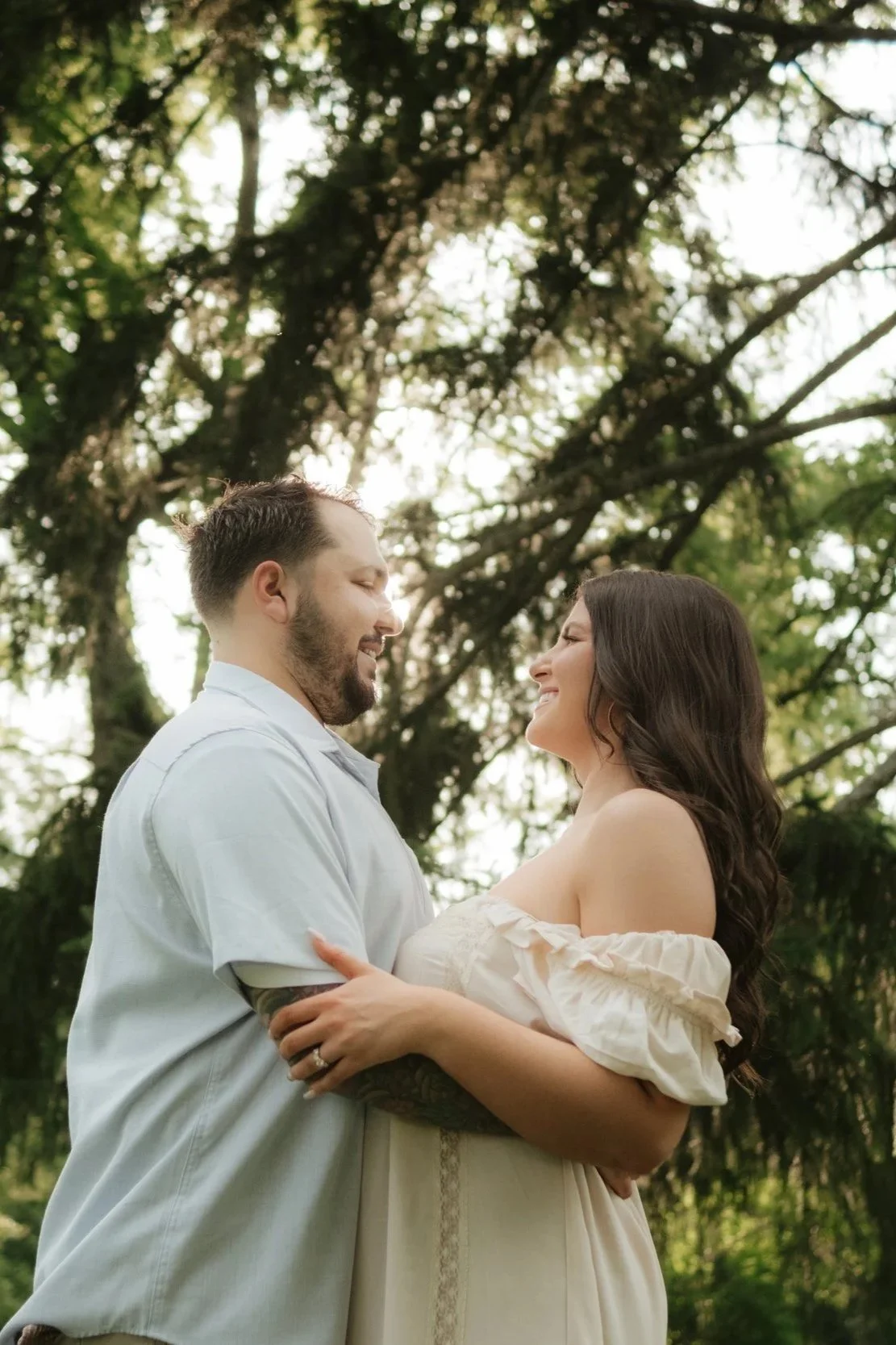A couple smiling and looking at each other in an outdoor setting with trees in the background.