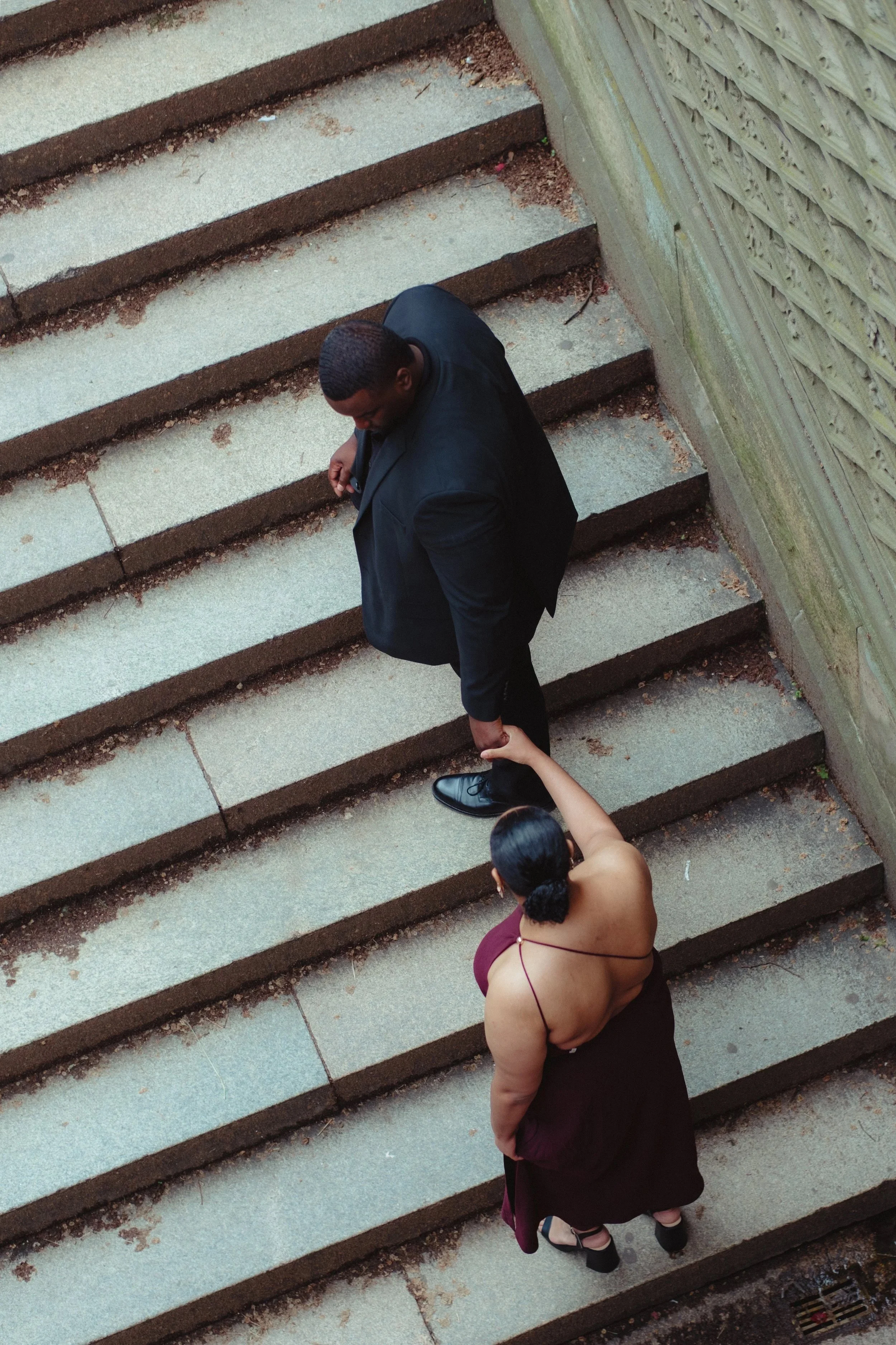 A man in a black suit and a woman in a burgundy dress with black heels are holding hands and descending outdoor concrete stairs. The woman is looking up at the man, who is looking down at her, with a textured wooden fence on the right side of the sta