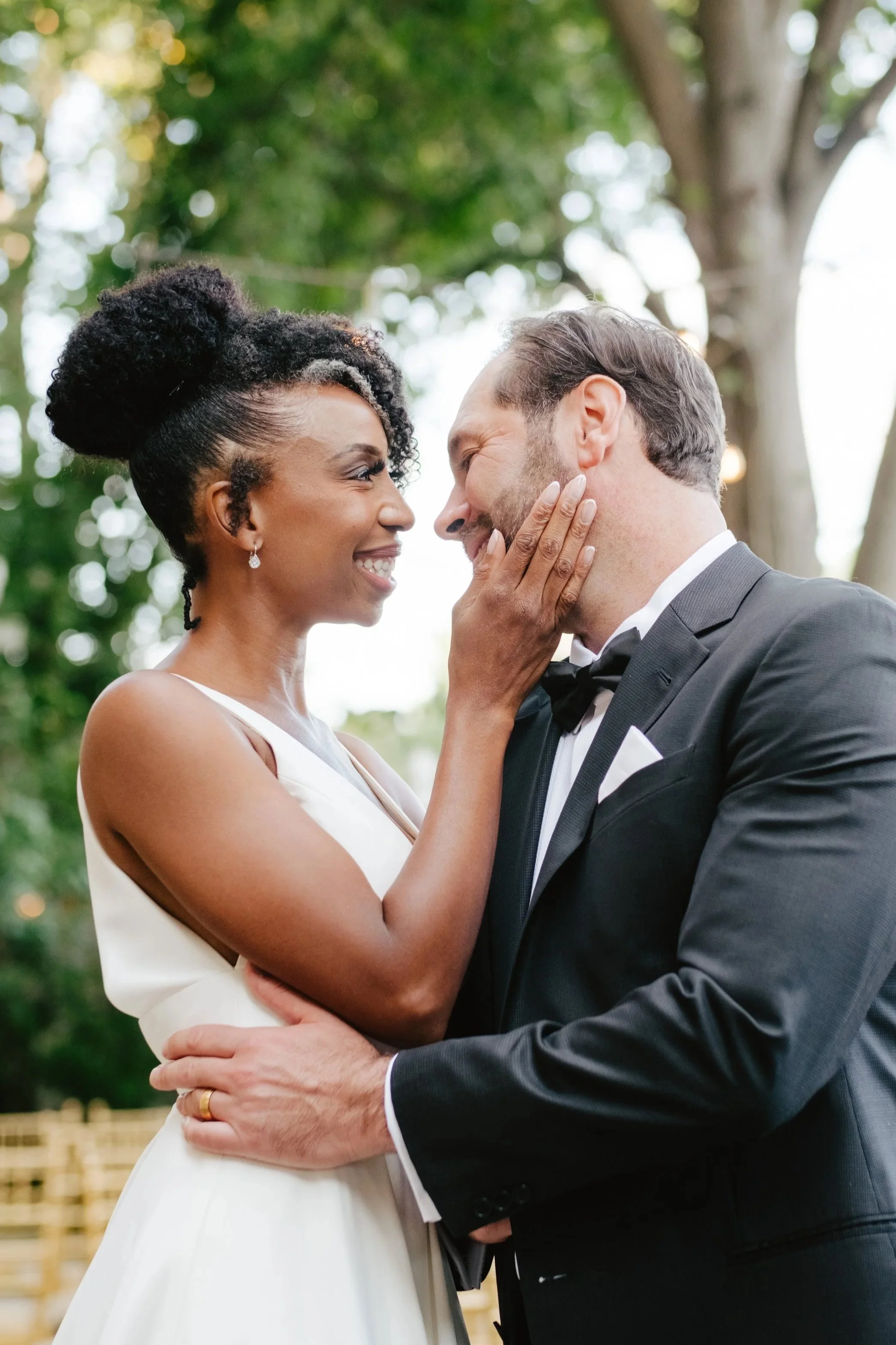 A bride and groom share an intimate moment outdoors, with the bride holding the groom's face and smiling at him as they look into each other's eyes. The bride is wearing a white wedding dress and the groom is in a black tuxedo.