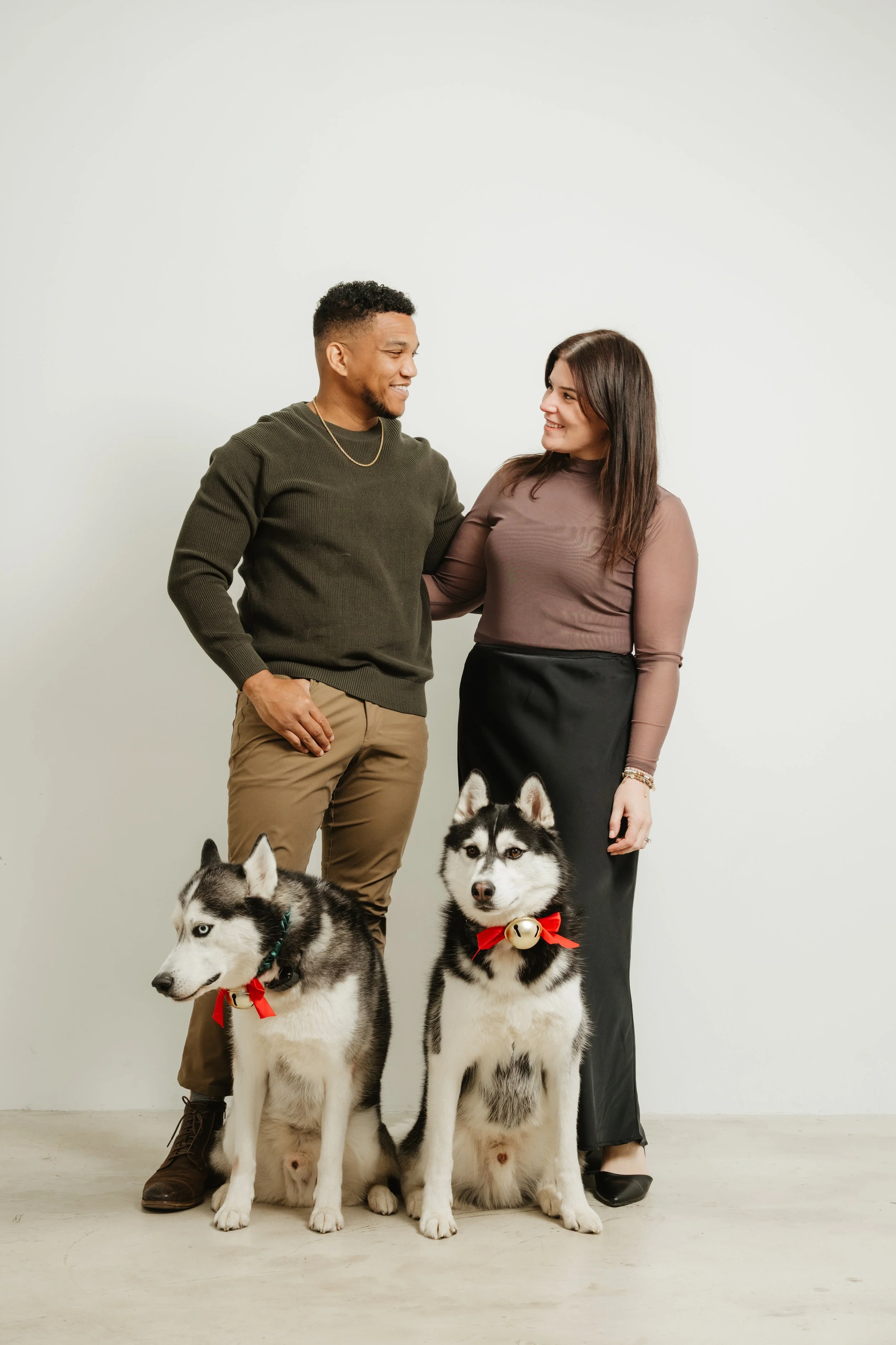 A man and woman standing together with two Siberian Huskies in front, the dogs wearing red bows with bells, in front of a plain white wall.