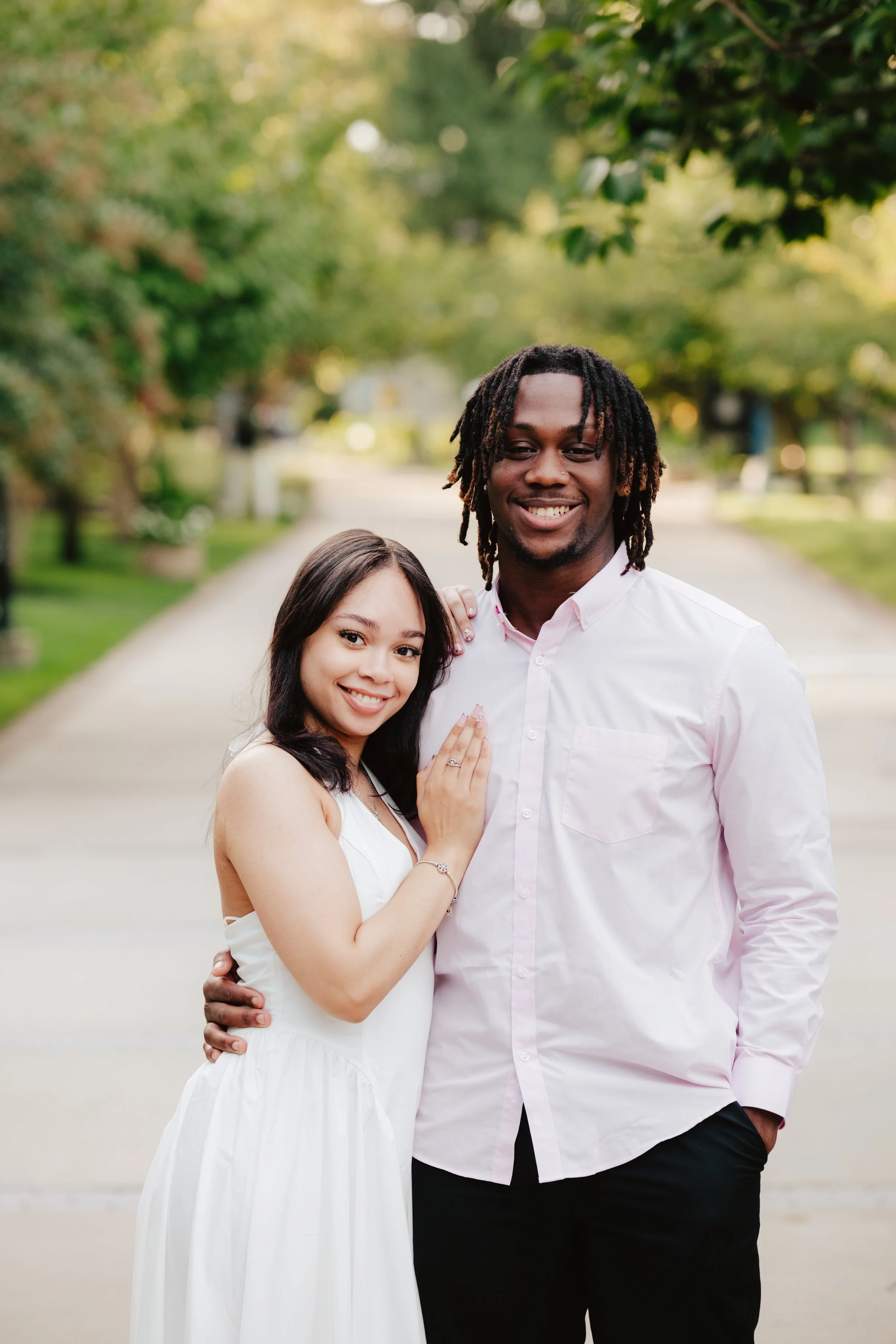 A young woman and a young man smiling and standing close together outdoors on a park path with green trees in the background. The woman has long dark hair and wears a white dress, while the man has dreadlocks and wears a light pink button-up shirt.