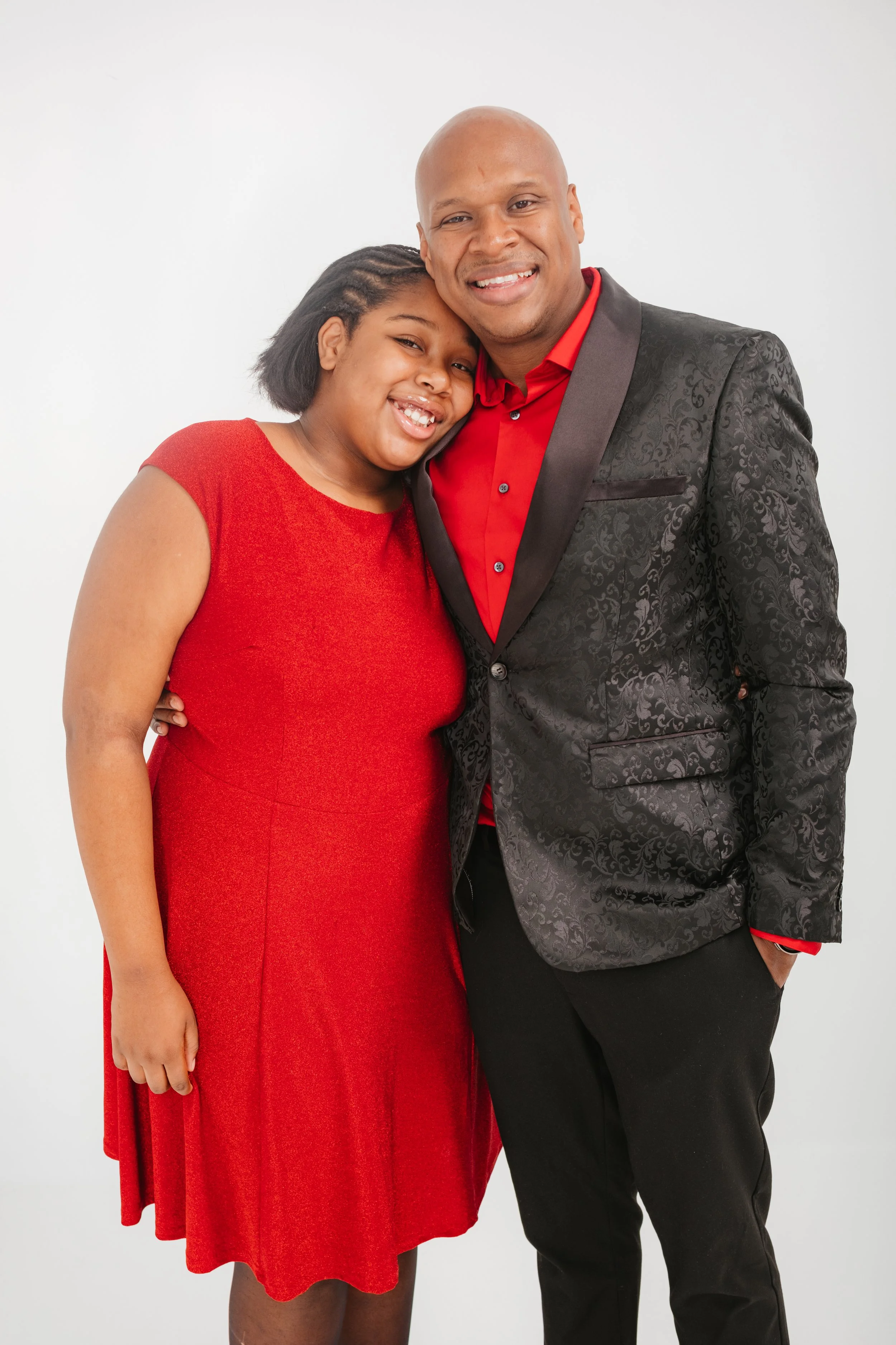 A smiling man and woman embracing each other, both dressed in red and black. The woman wears a red dress and the man a red shirt with a black blazer, standing against a plain white background.