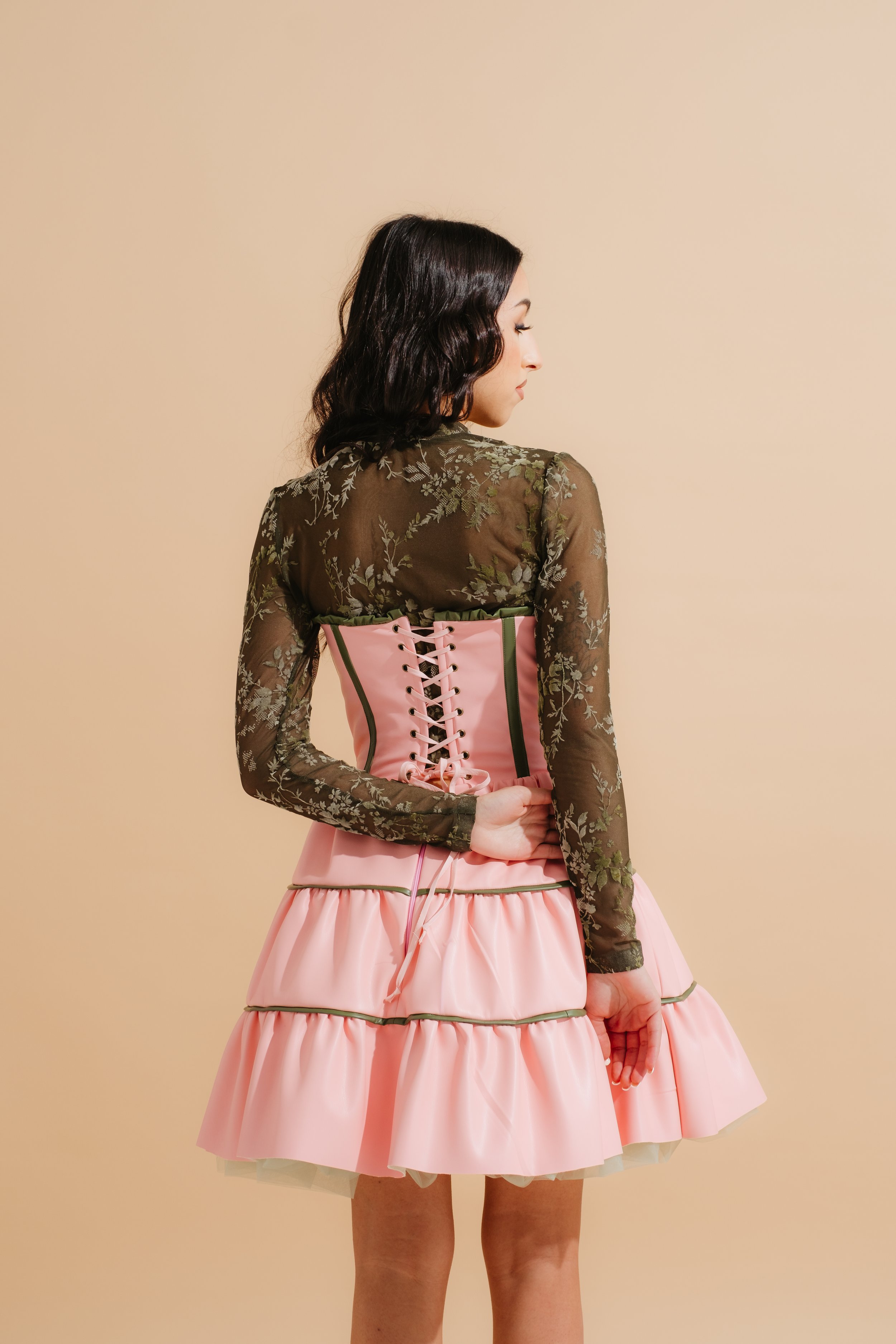 A woman with dark, wavy hair in a side profile facing left, wearing a sheer olive green top with floral patterns and a pink corset with lace-up detail, over a pink ruffled skirt, against a plain beige background.