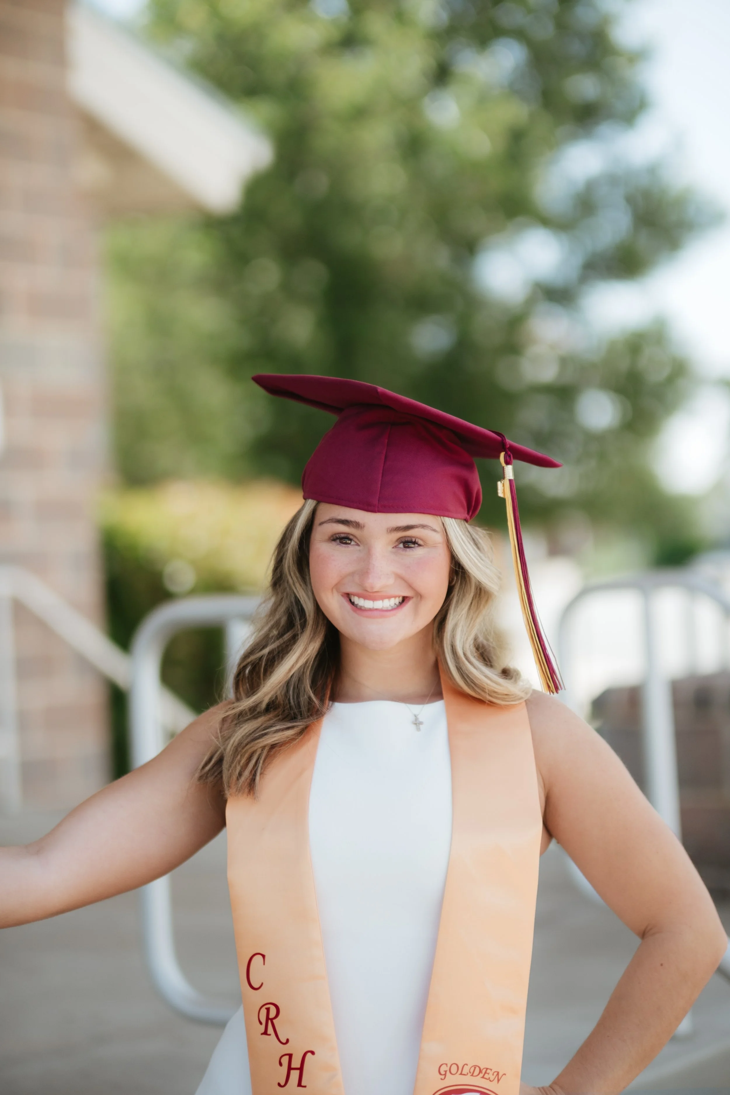 Young woman in a white dress wearing a maroon graduation cap and a light peach sash with red and gold lettering, smiling outdoors with blurred greenery and brick steps in the background.