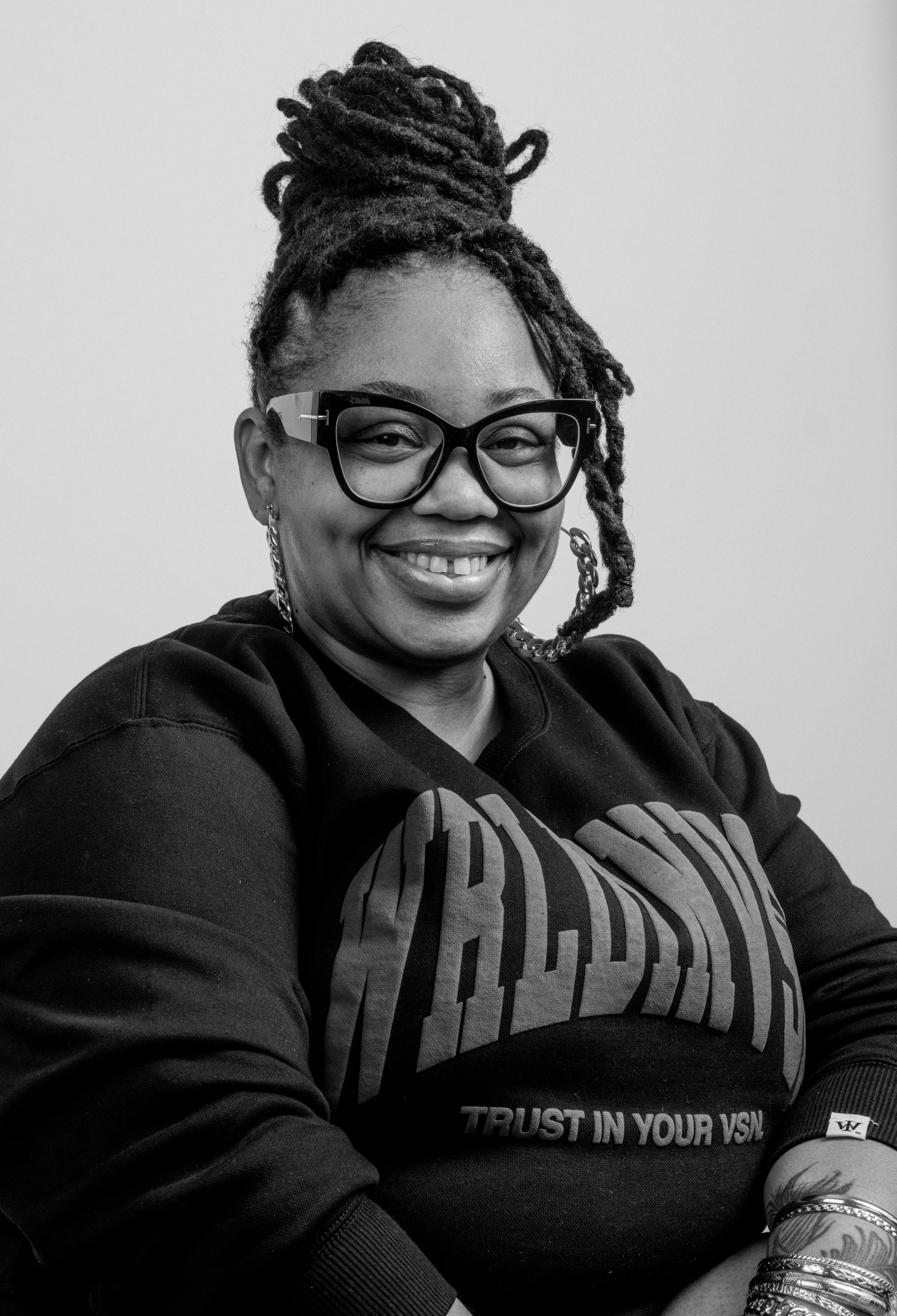 A smiling woman with glasses and dreadlocks styled in a bun, wearing a black sweatshirt with text, and accessorized with earrings and bracelets, posed against a plain background.