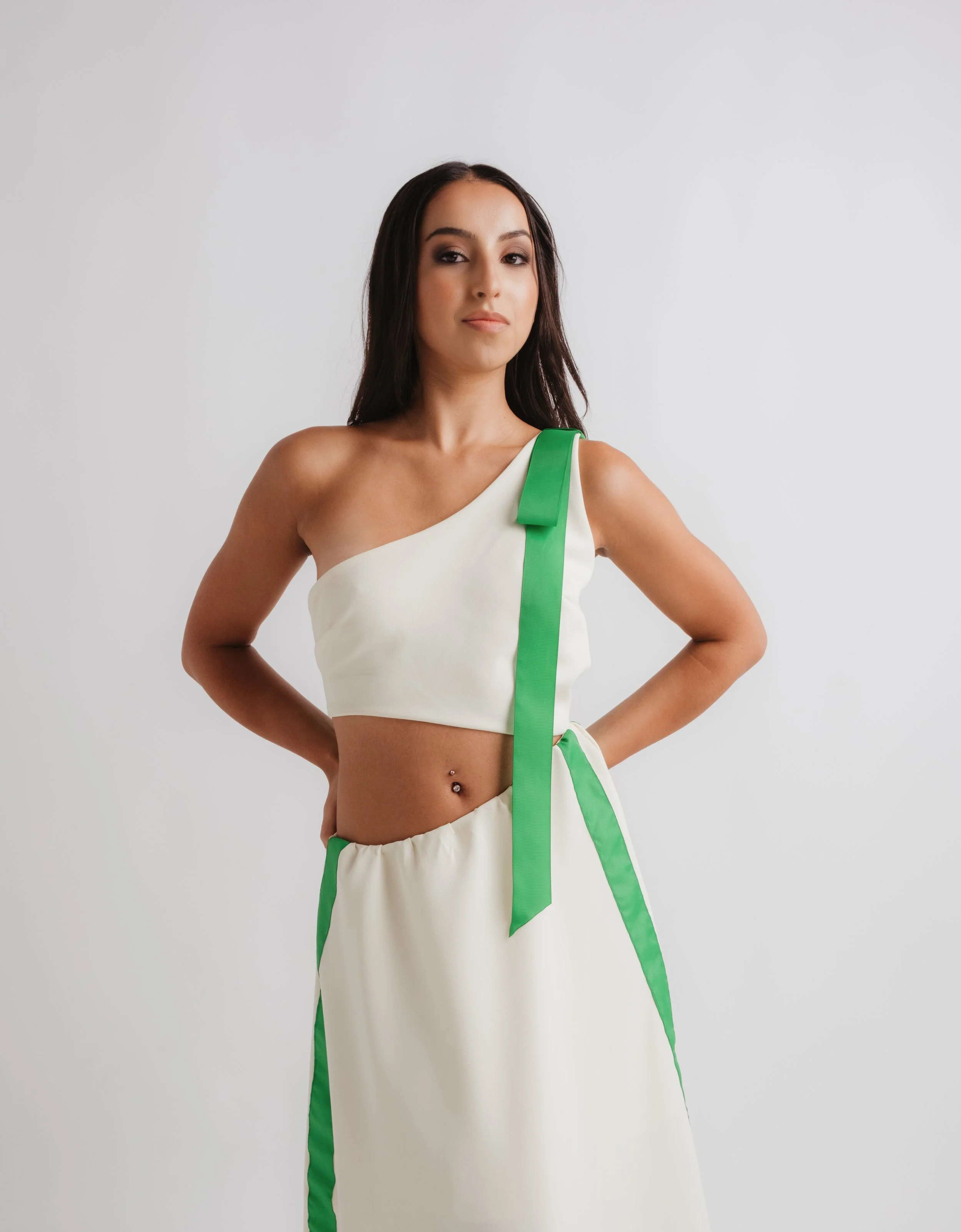 Young woman with long dark hair wearing a cream-colored crop top with green accents and matching cream-colored skirt with green stripes, standing against a plain white wall.