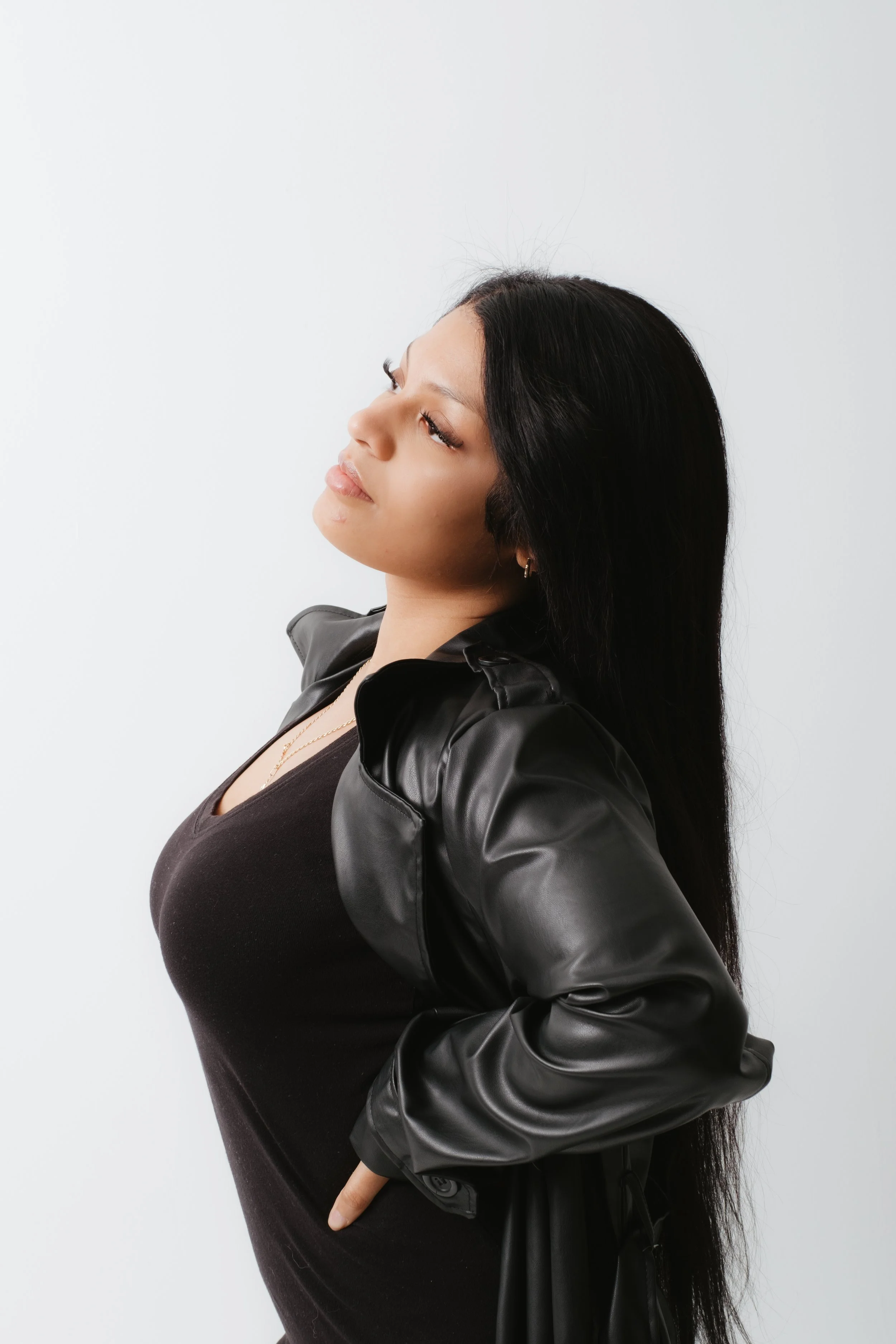 Young woman with long black hair wearing a black leather jacket and dark top, posing with her hands on her hips against a plain white background.
