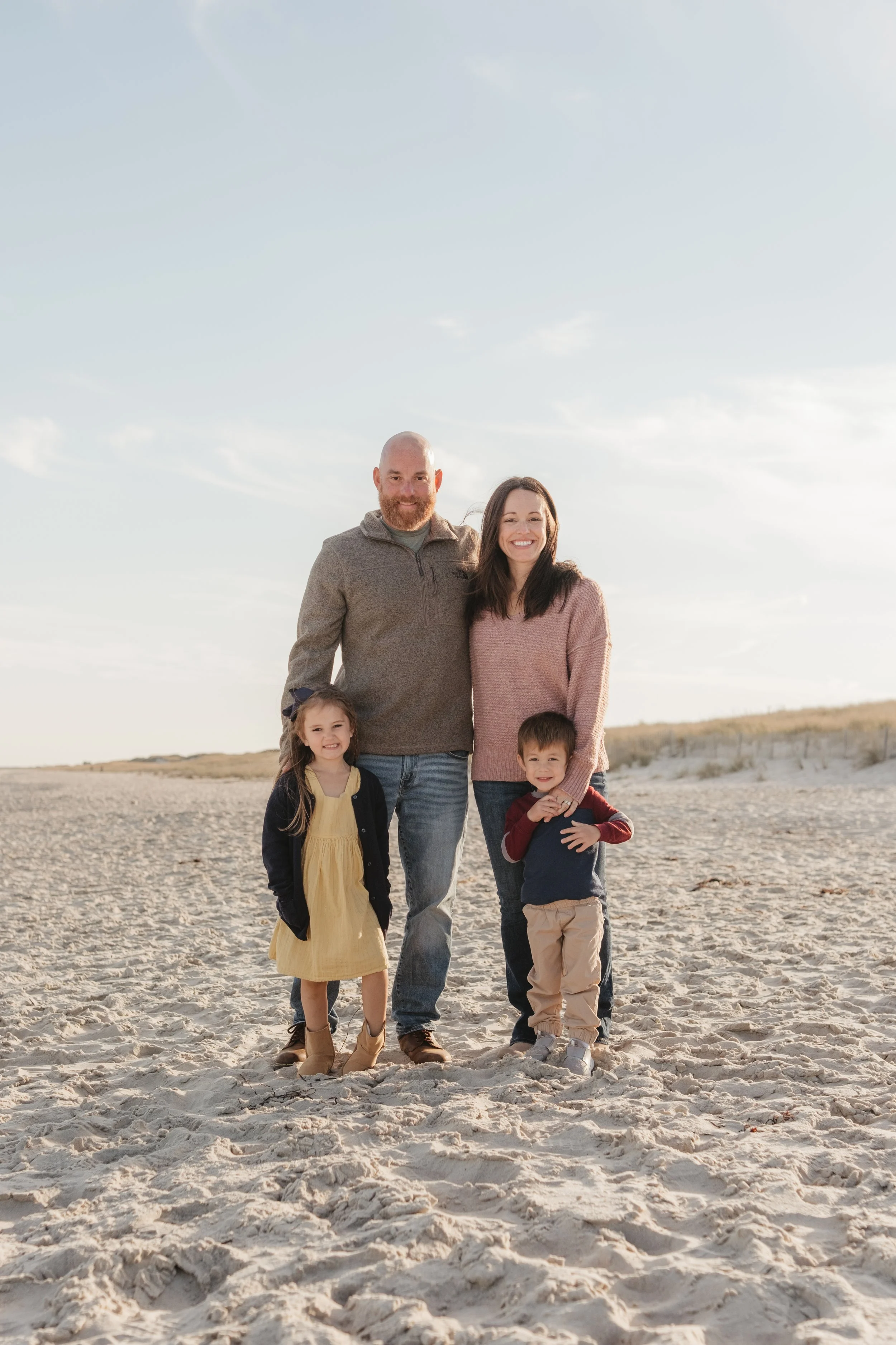 A family of four standing on the sand at the beach, smiling at the camera under a partly cloudy sky.