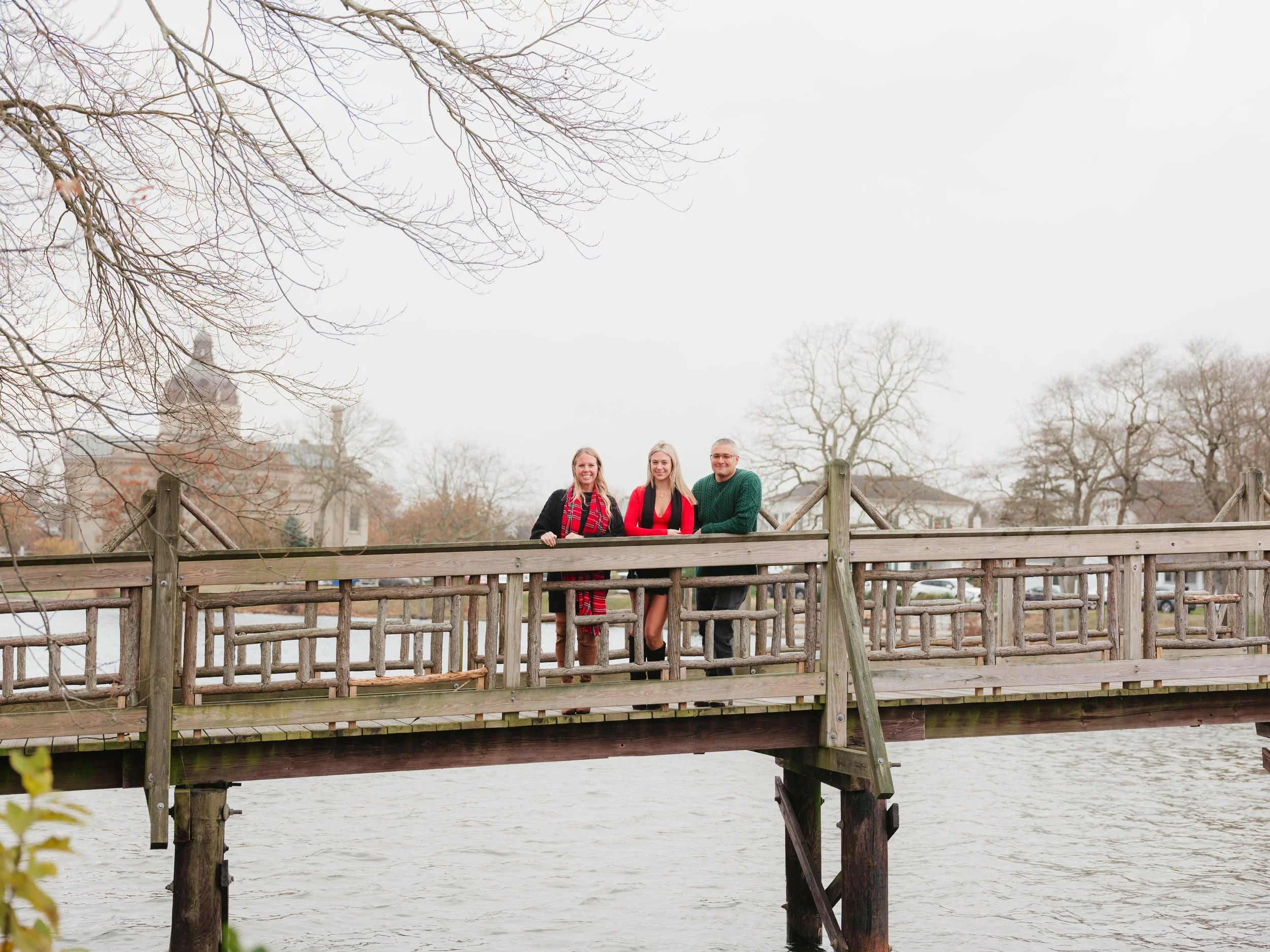 Three people standing on a wooden bridge over water, with leafless trees and buildings in the background, during overcast weather.