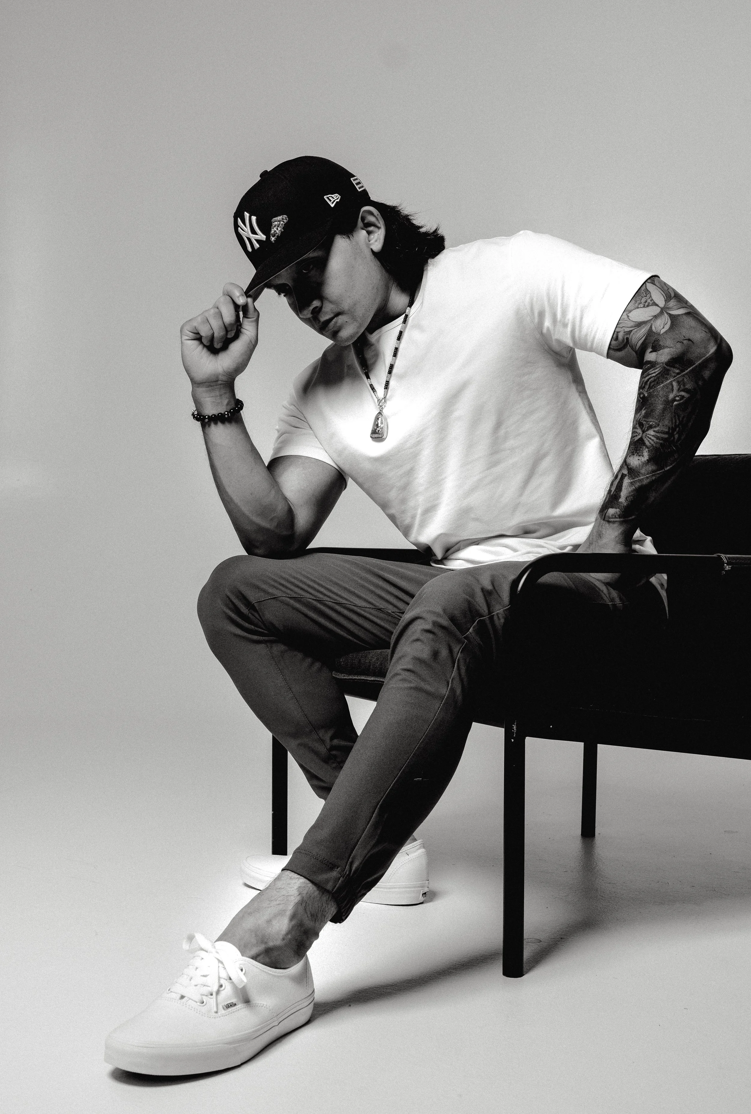 A young man wearing a New York Yankees cap, a white t-shirt, gray pants, and white sneakers, sitting on a black chair, touching the brim of his cap with one hand and resting his other hand on his hip, with tattoos on his left arm, wearing a necklace 