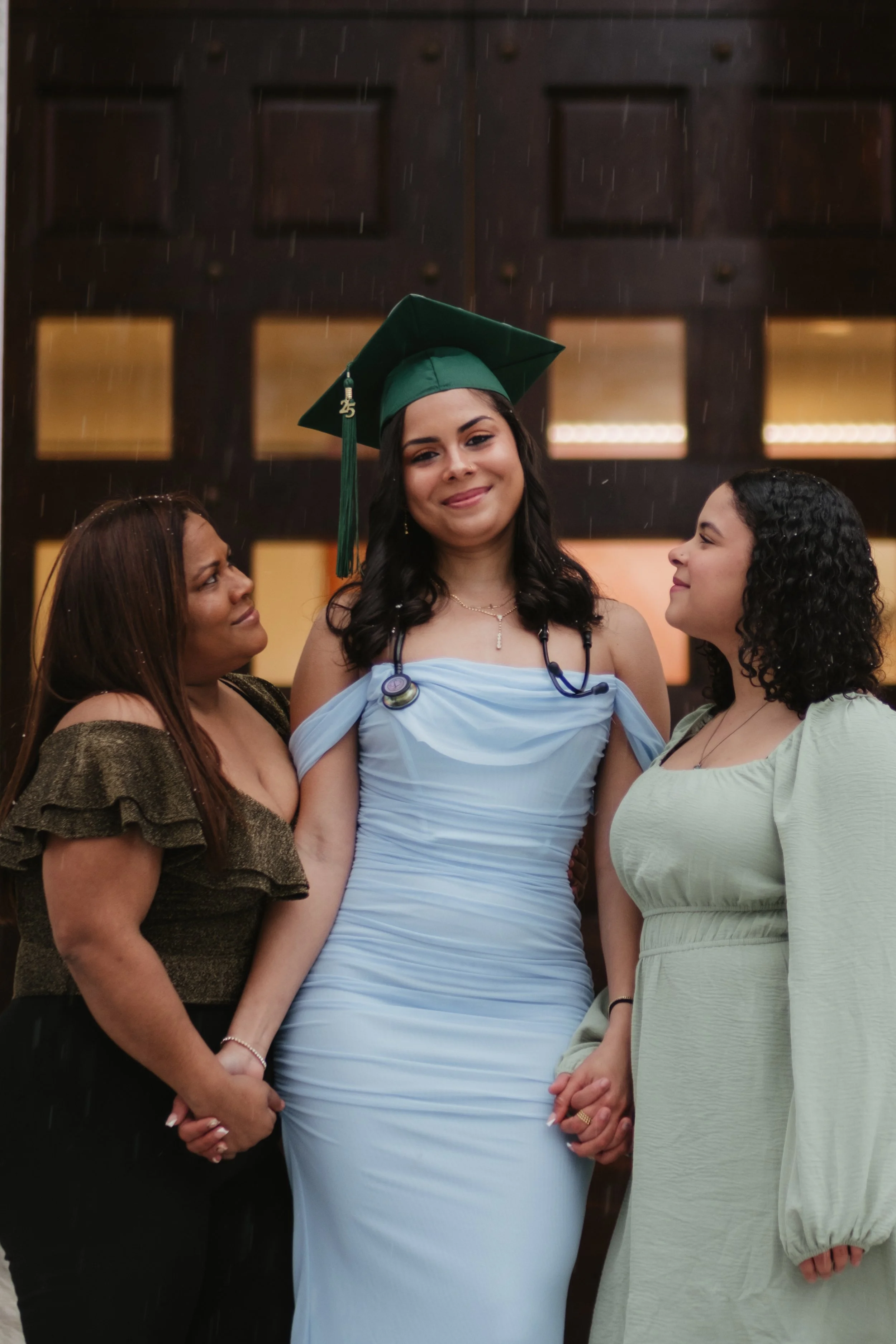 A young woman in a light blue gown and green graduation cap with a tassel stands between two women, holding hands, outside in the rain at night. The women are smiling and looking at each other.