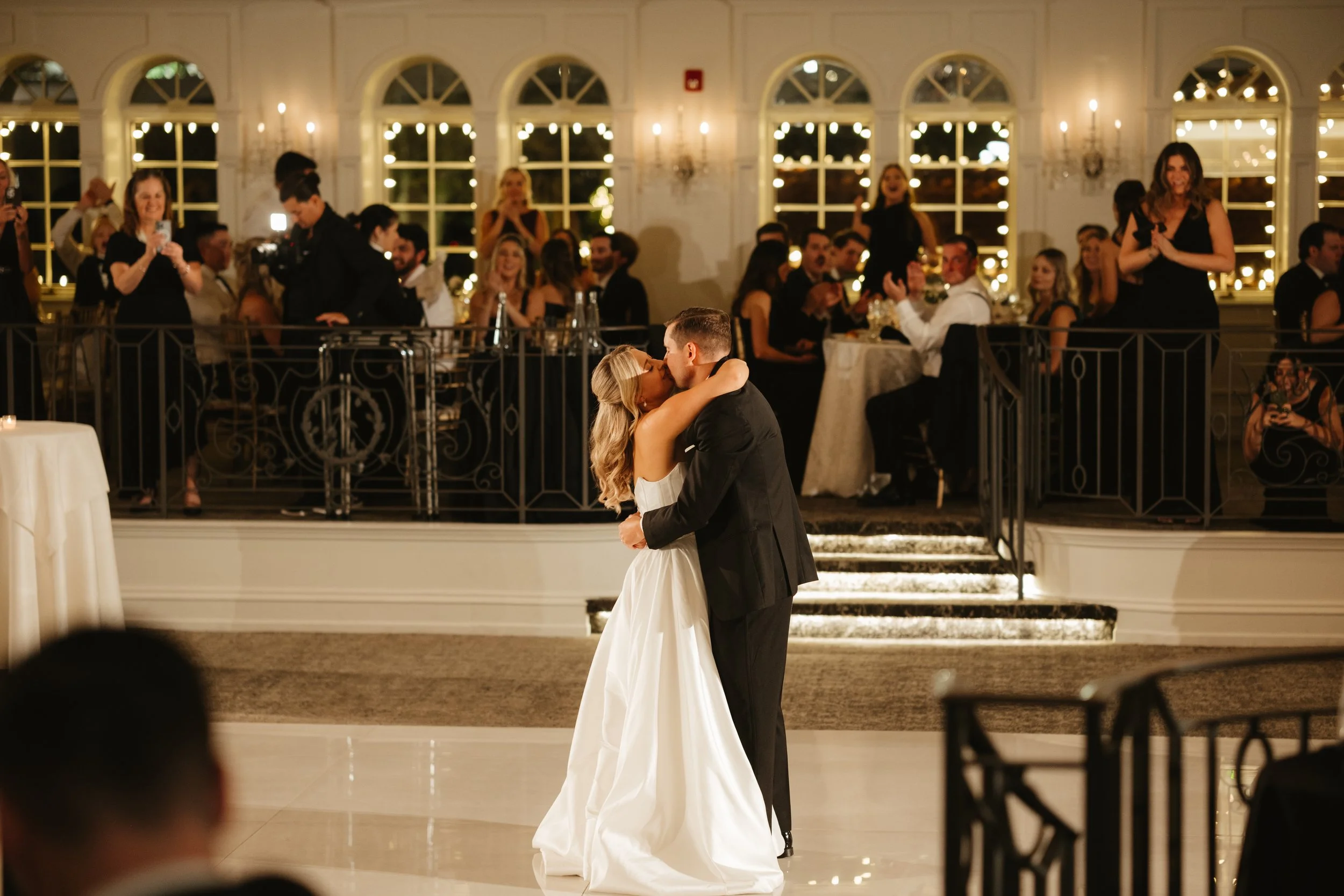 A bride and groom share their first dance at their wedding reception, surrounded by seated guests and illuminated by warm lighting.