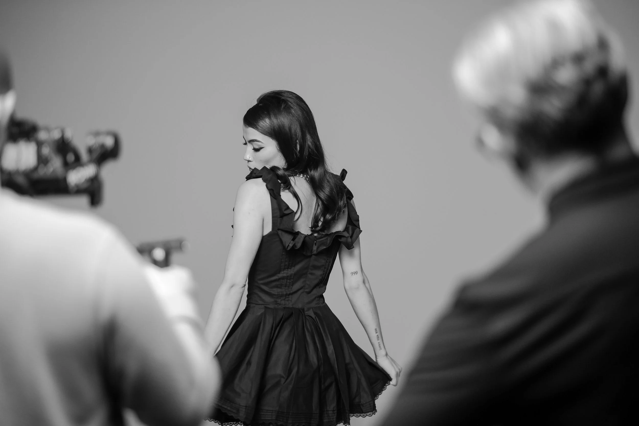 Black and white photo of a woman in a dress, looking over her shoulder, while two people take pictures or film her.