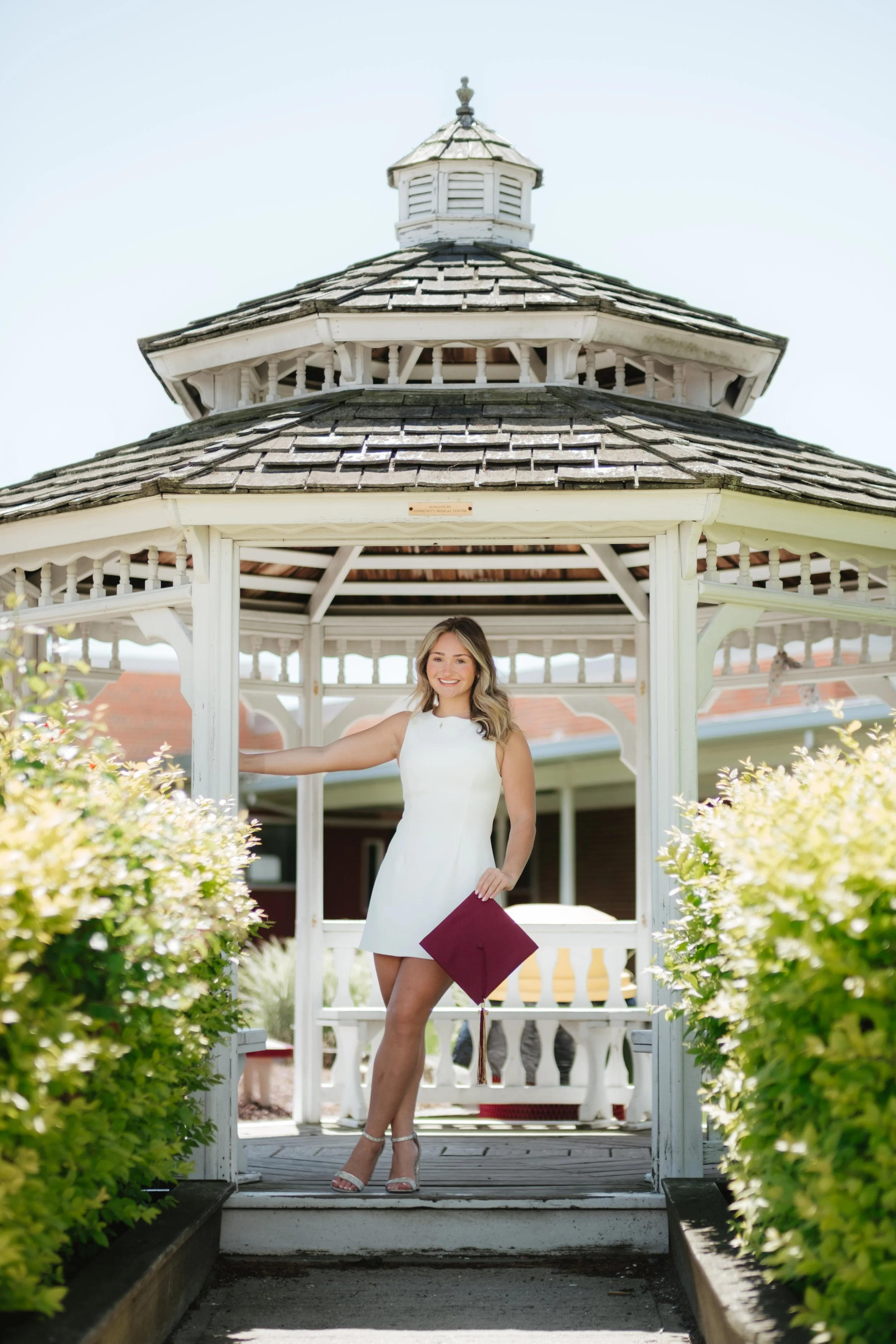 A woman in a white dress holding a maroon graduation cap standing inside a white gazebo outdoors with blooming bushes and a red-roofed building in the background.