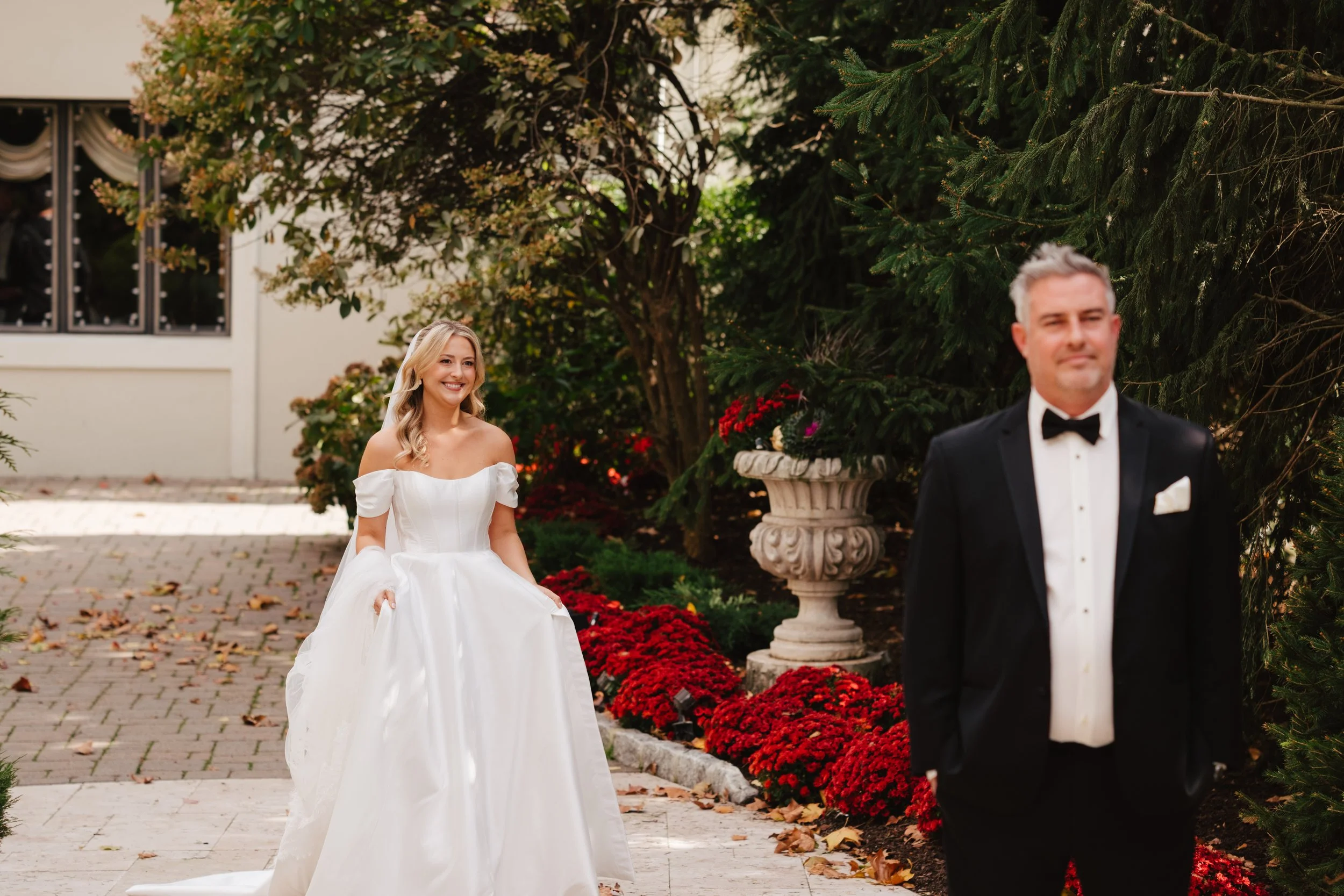 A bride in a white dress with puffed sleeves smiling as she approaches a groom in a black tuxedo outdoors near a garden with red flowers and large green trees.