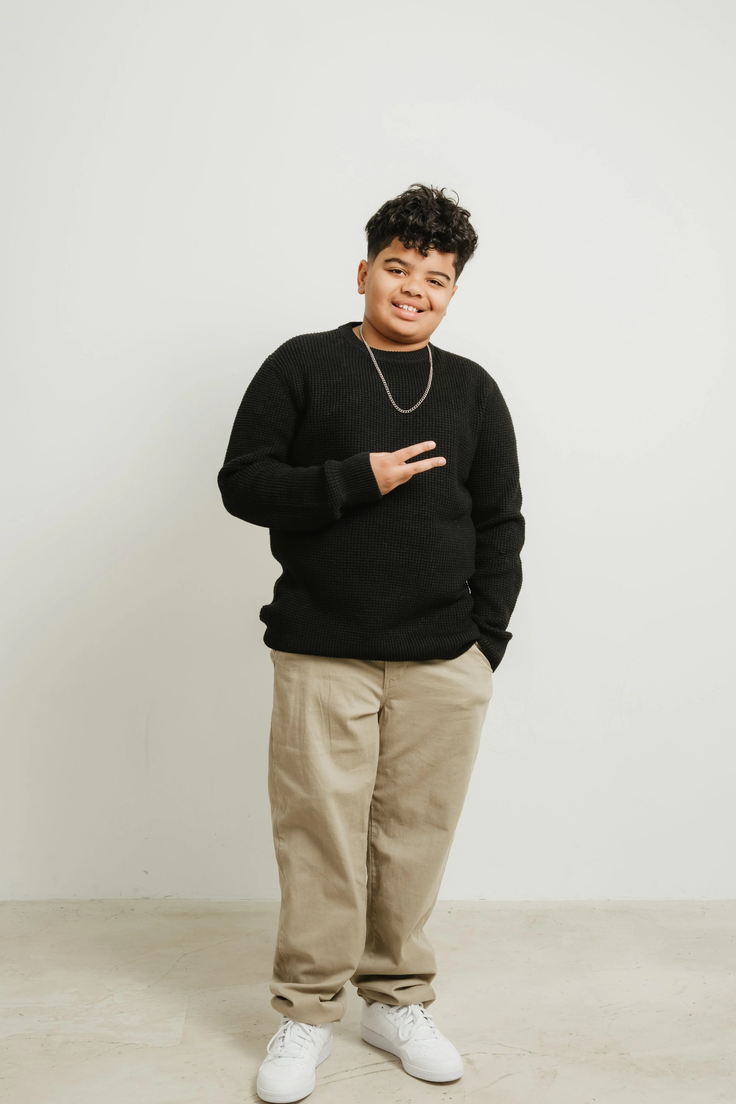 A young man with curly hair smiling, wearing a black sweater, beige pants, white sneakers, and a silver chain, standing against a plain white wall.
