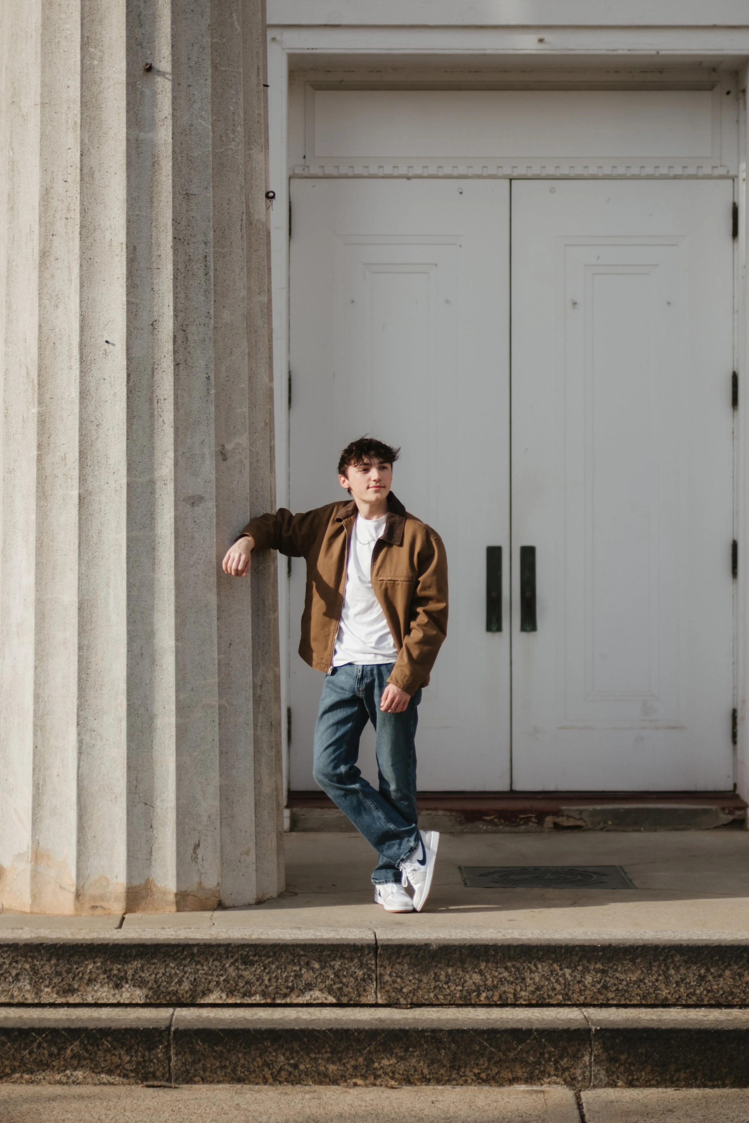 A young man standing outside on a sidewalk, leaning against a concrete column with a large white door behind him.