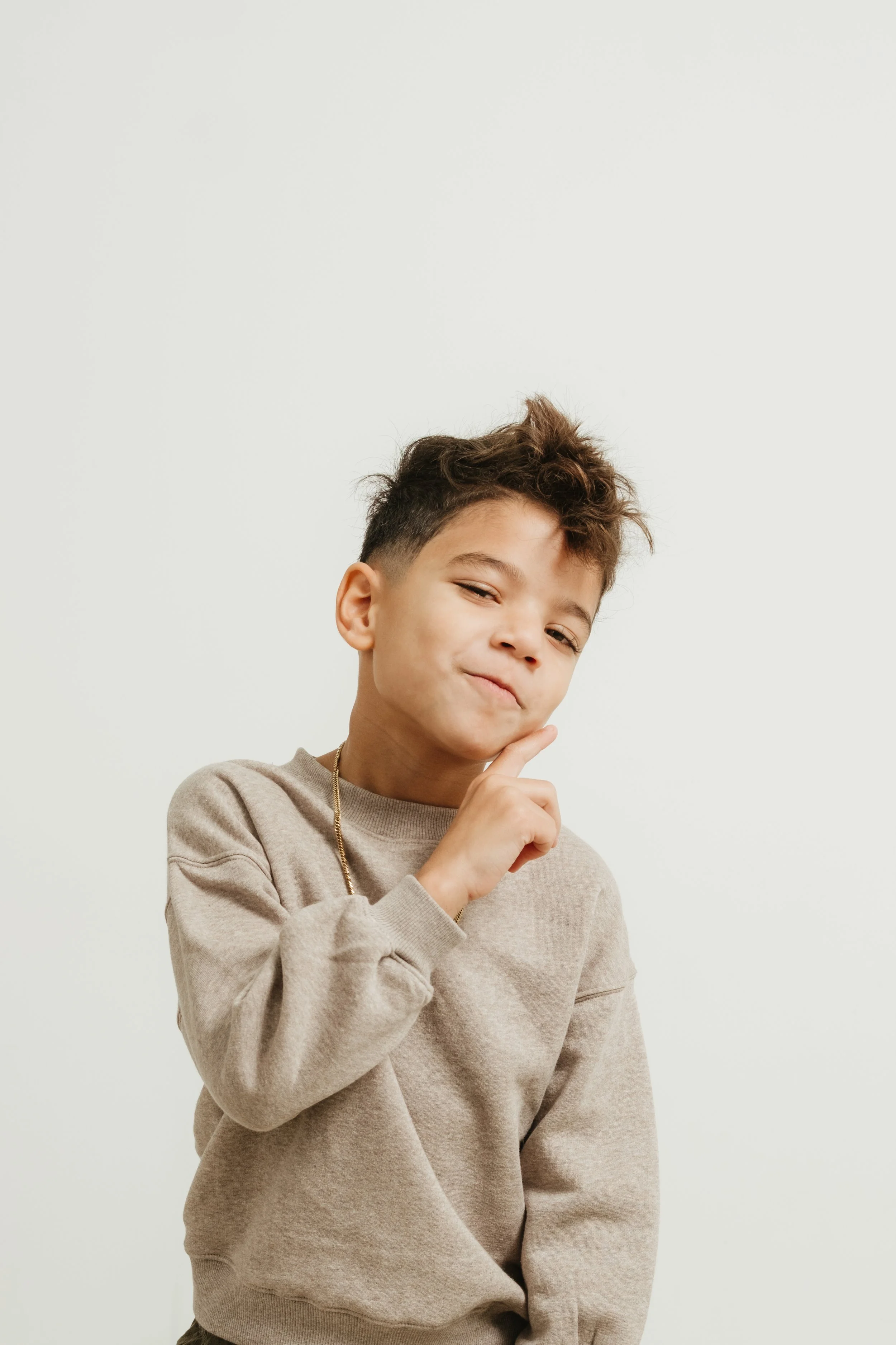 A young boy with a playful expression, wearing a beige sweatshirt and a gold chain, poses with one finger on his chin against a plain white background.