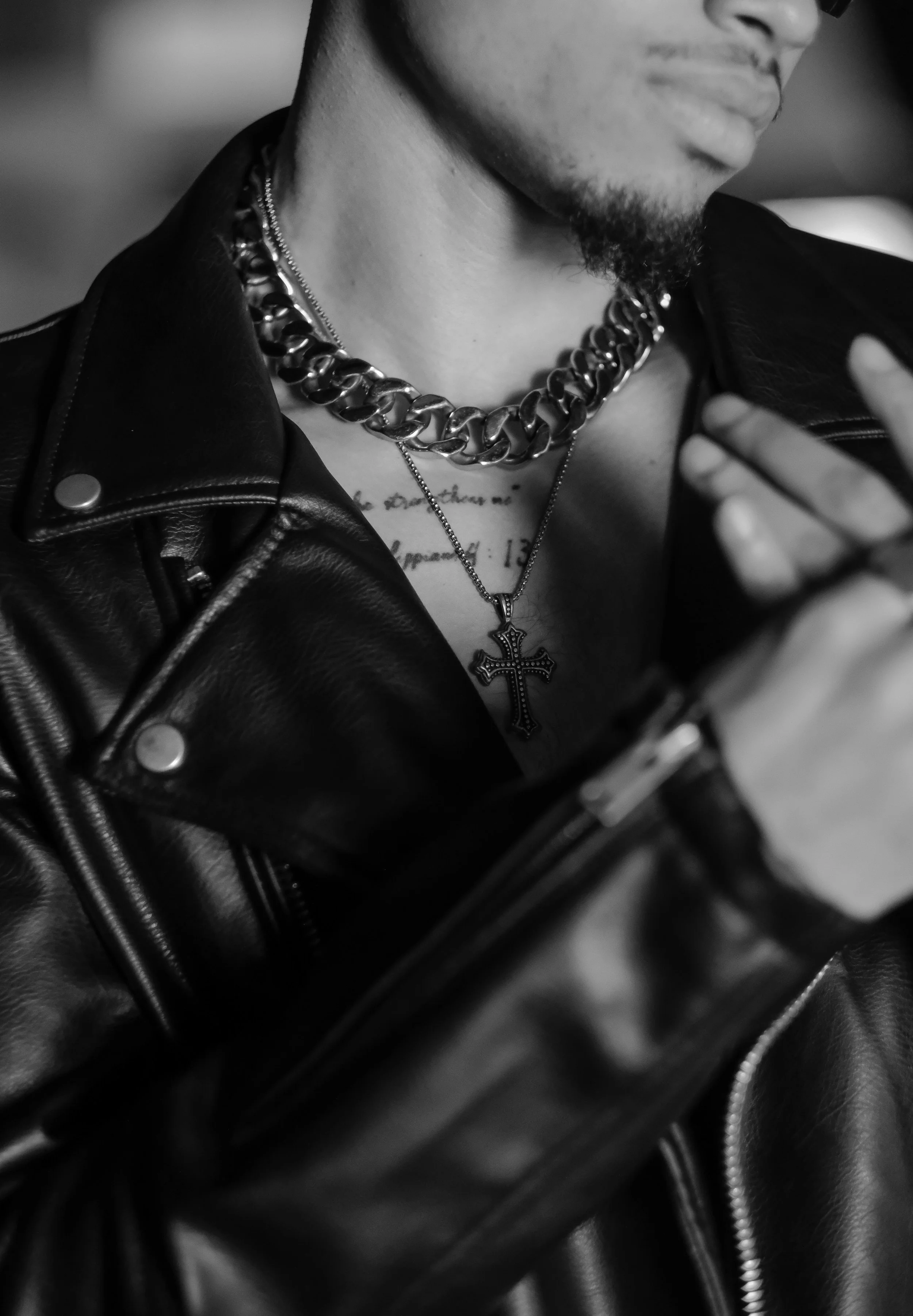 A close-up black and white photo of a person wearing a leather jacket, a chunky chain necklace, and a necklace with a cross pendant. The person's face is partially visible, showing their chin and lips.