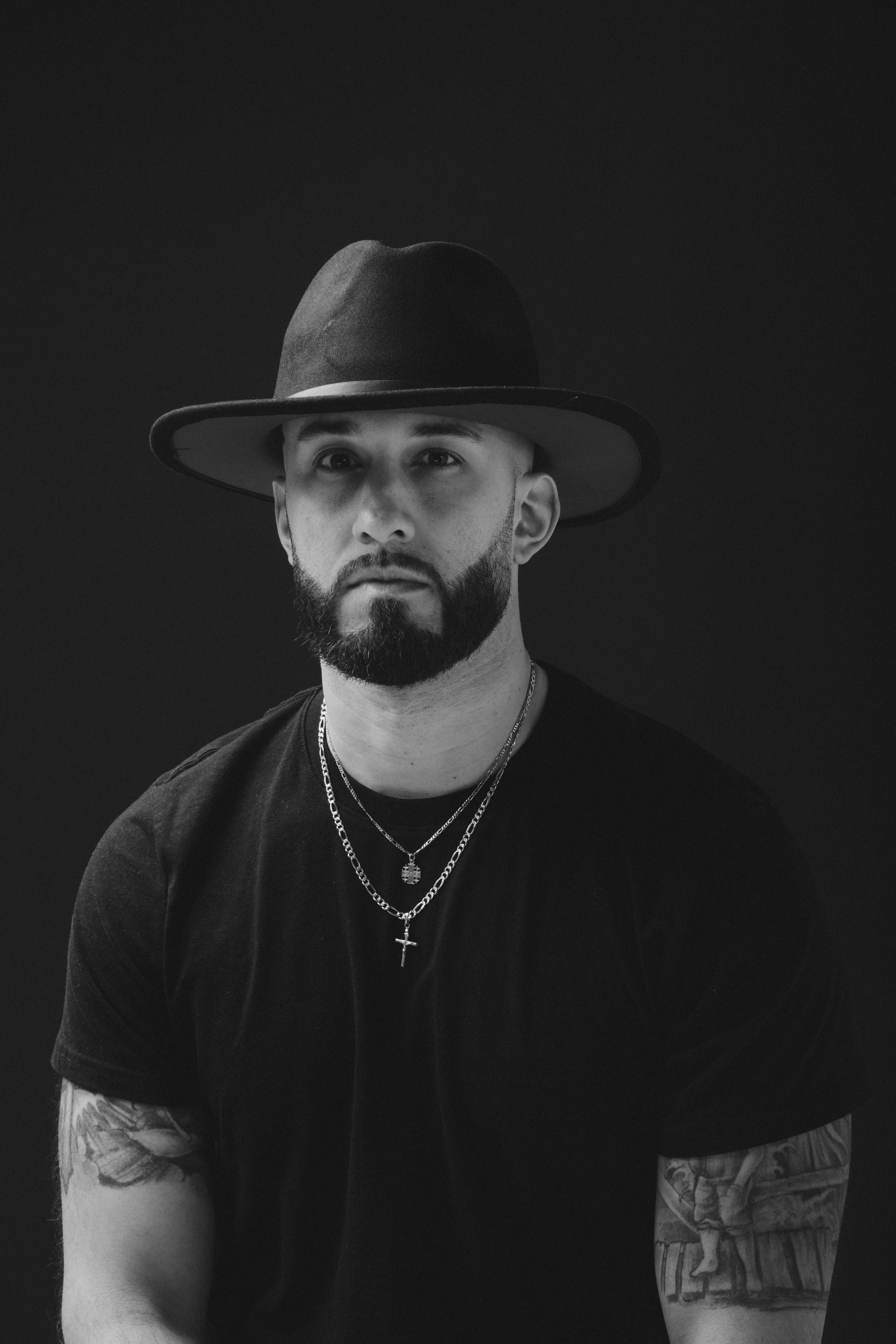 Black and white portrait of a man with a beard, wearing a wide-brimmed hat, black t-shirt, and layered necklaces with crosses and a gemstone, against a dark background.