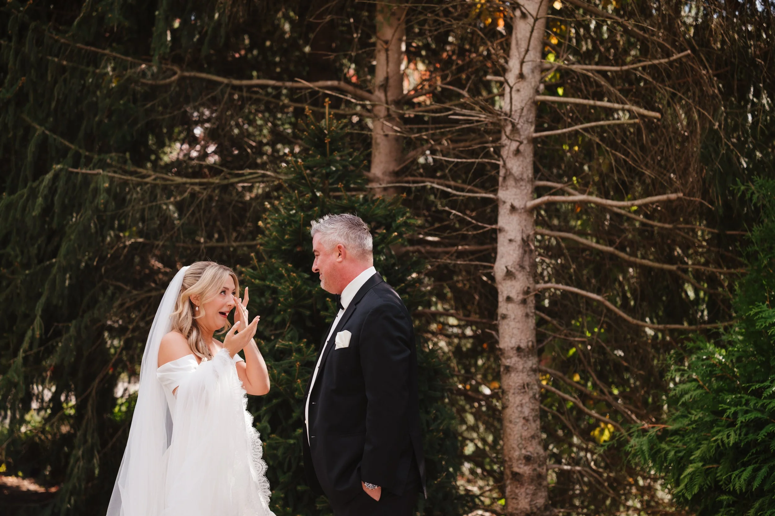 A bride and groom standing outdoors in front of a forest with trees, during a wedding ceremony, emotional moment as the bride reacts with surprise or joy.