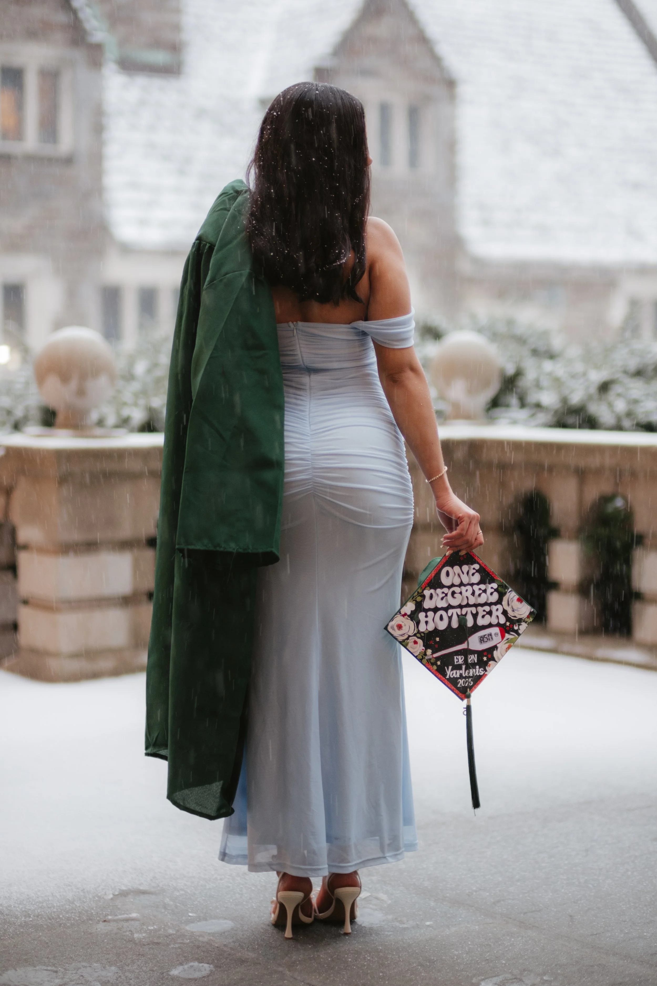 A woman in a white dress holding a graduation cap that says 'One Degree Hotter', standing on a balcony in the rain, with buildings in the background.