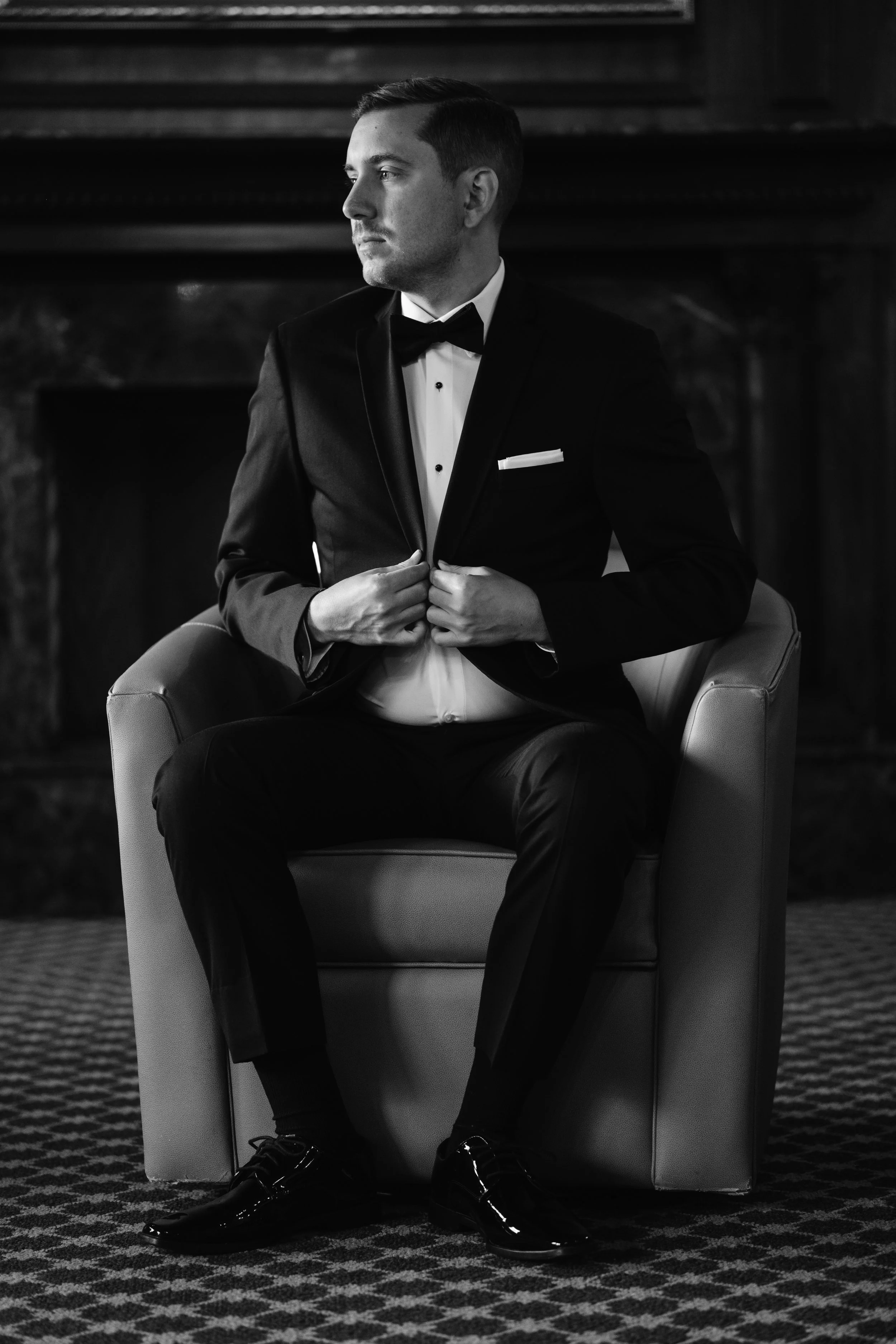 Black and white photo of a man in a tuxedo sitting on a chair, adjusting his jacket, in a formal setting.